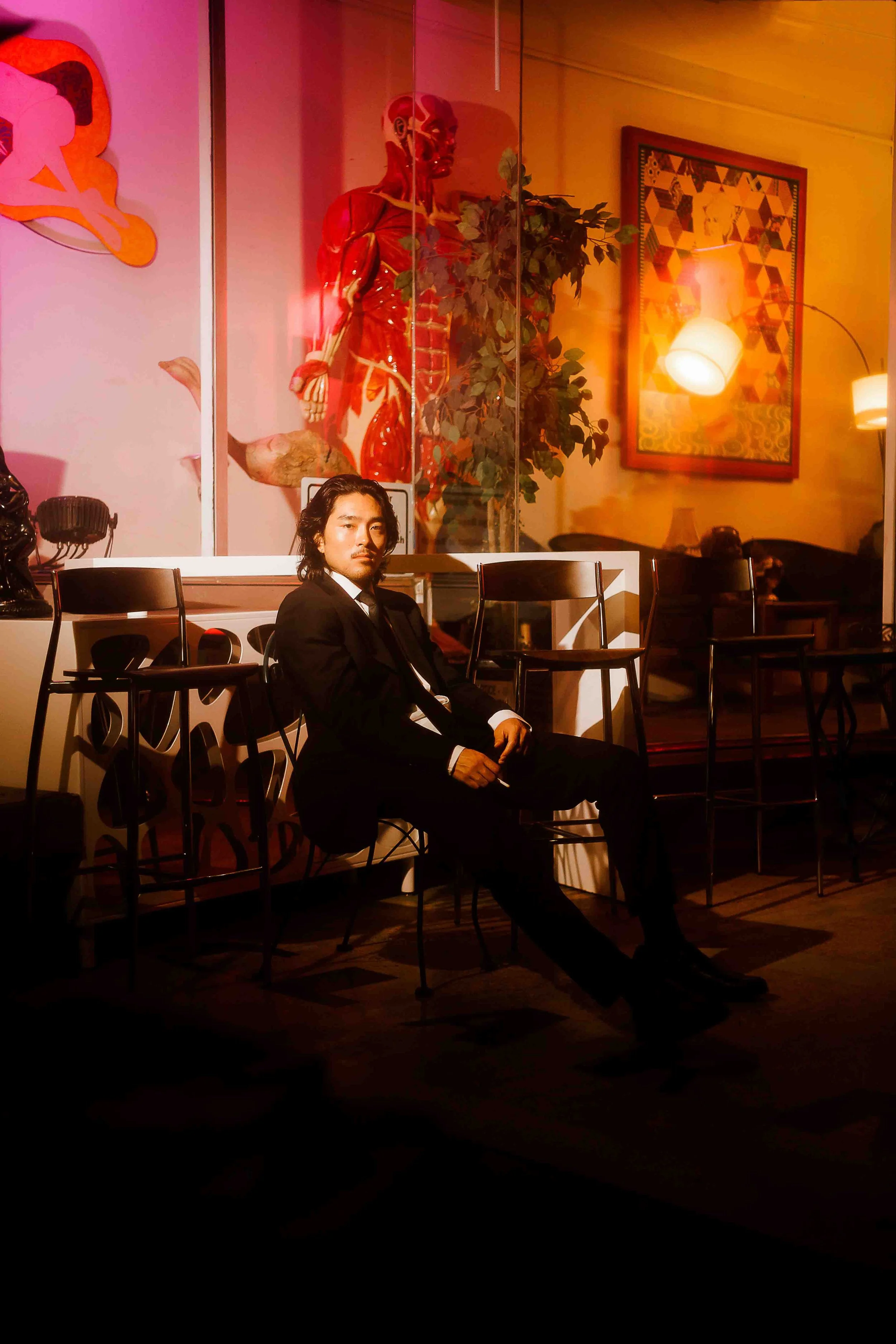 Houston editorial portrait of a man in a black suit sitting on a chair in a dimly lit modern room with artistic wall art and a large anatomical sculpture behind a glass partition, illuminated by warm ambient lighting