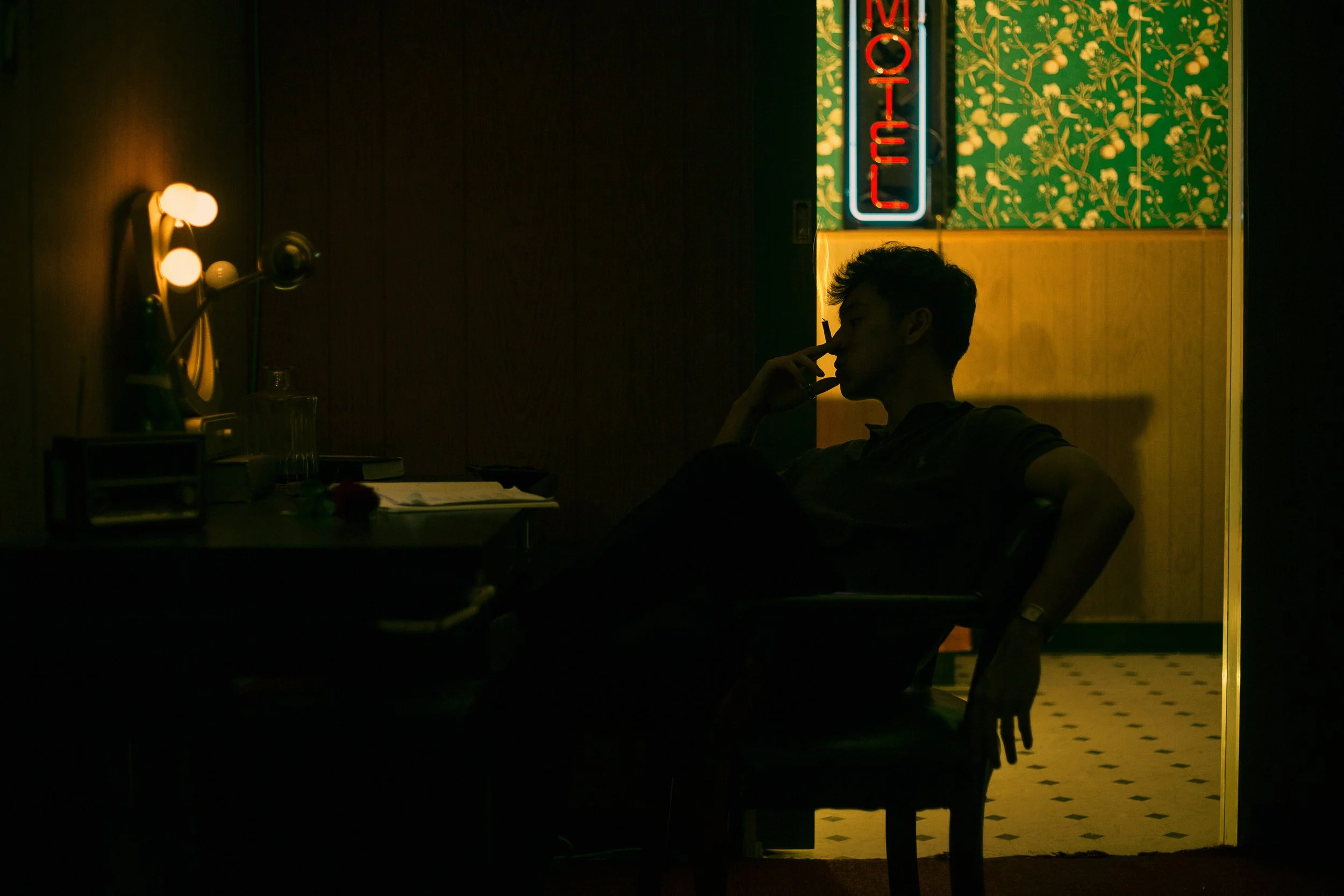 A person sitting in a dimly lit room, silhouette with a hand near their face, next to a table with books and glasses, a mirror with a lamp, and a neon sign that reads 'Open' in the background.