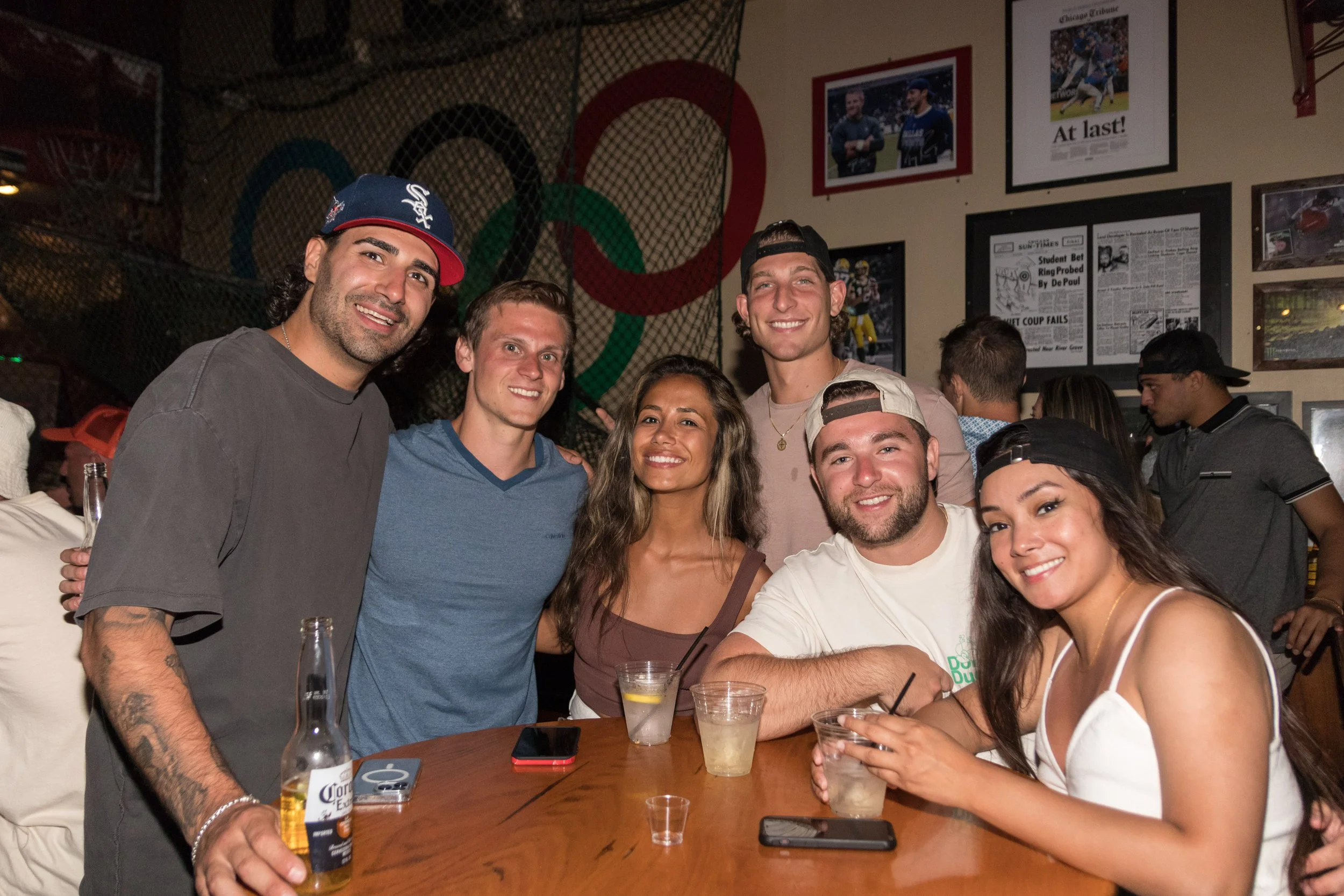 A group of six young adults smiling and posing for a photo at a bar or restaurant. They are sitting around a round table with drinks and phones. The background has sports memorabilia, posters, and a large Olympic rings display.