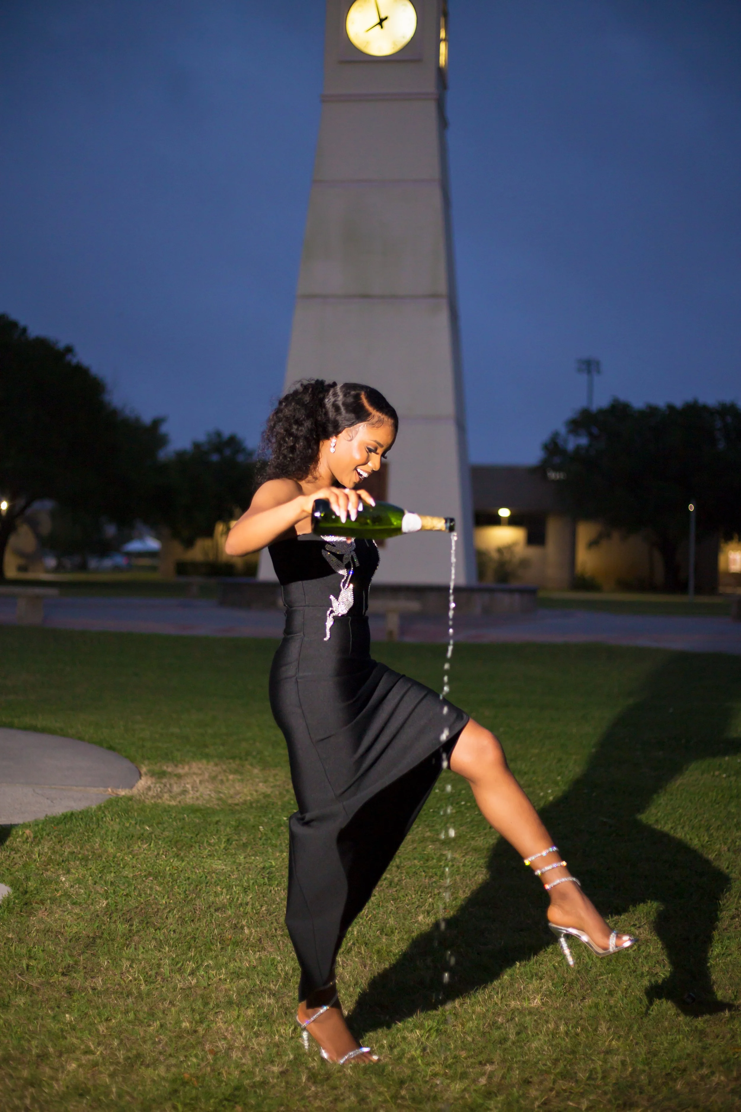 A woman in a black dress and high heels is pouring champagne on the grass in front of a clock tower at dusk.