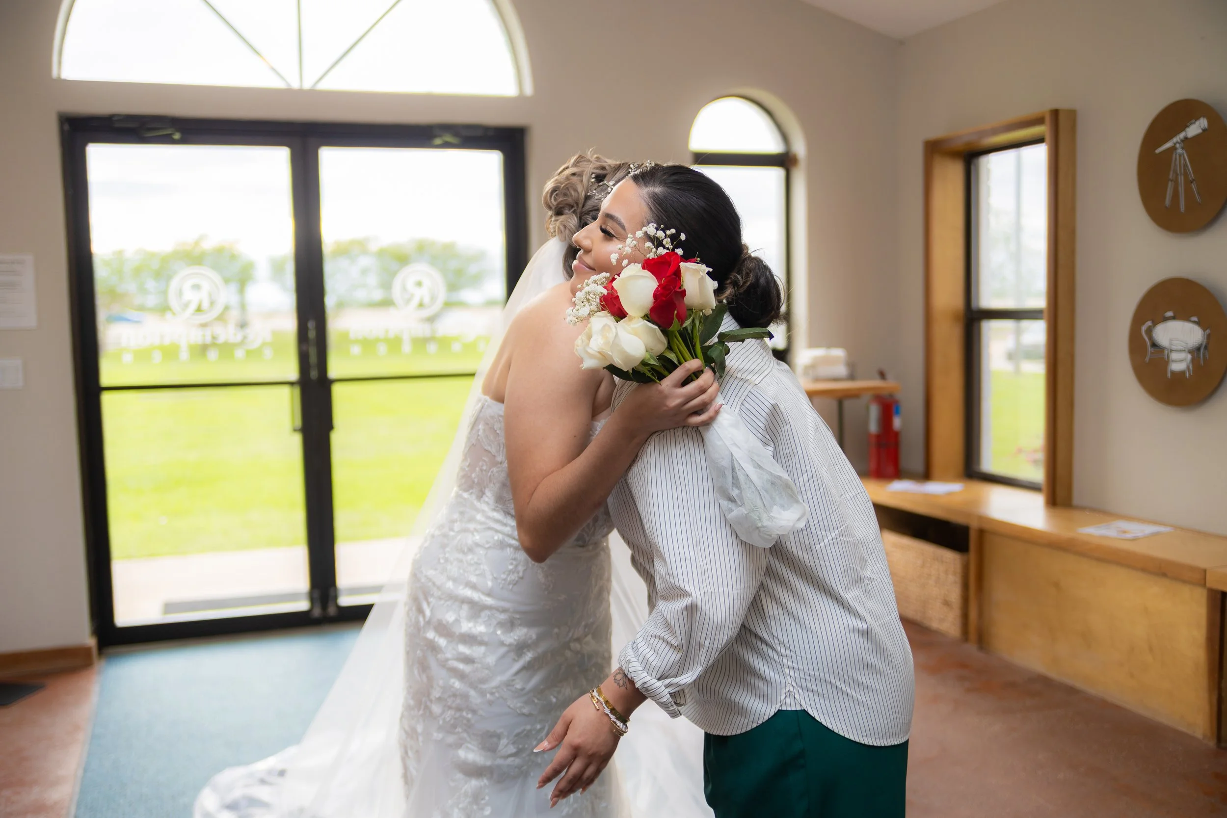 A bride and another woman hugging indoors, with the bride holding a bouquet of red and white roses.