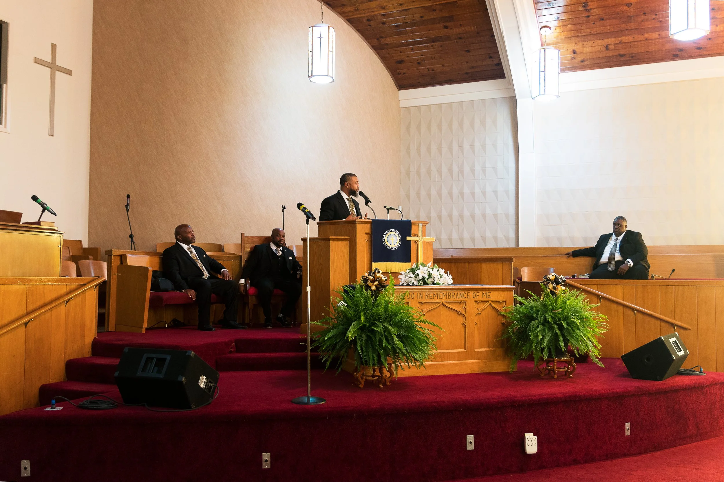 A church service with a preacher and three men seated on the stage. The preacher is standing at a pulpit, speaking into a microphone. The stage is decorated with flowers and greenery, and there are two large speakers on the floor. The church interior