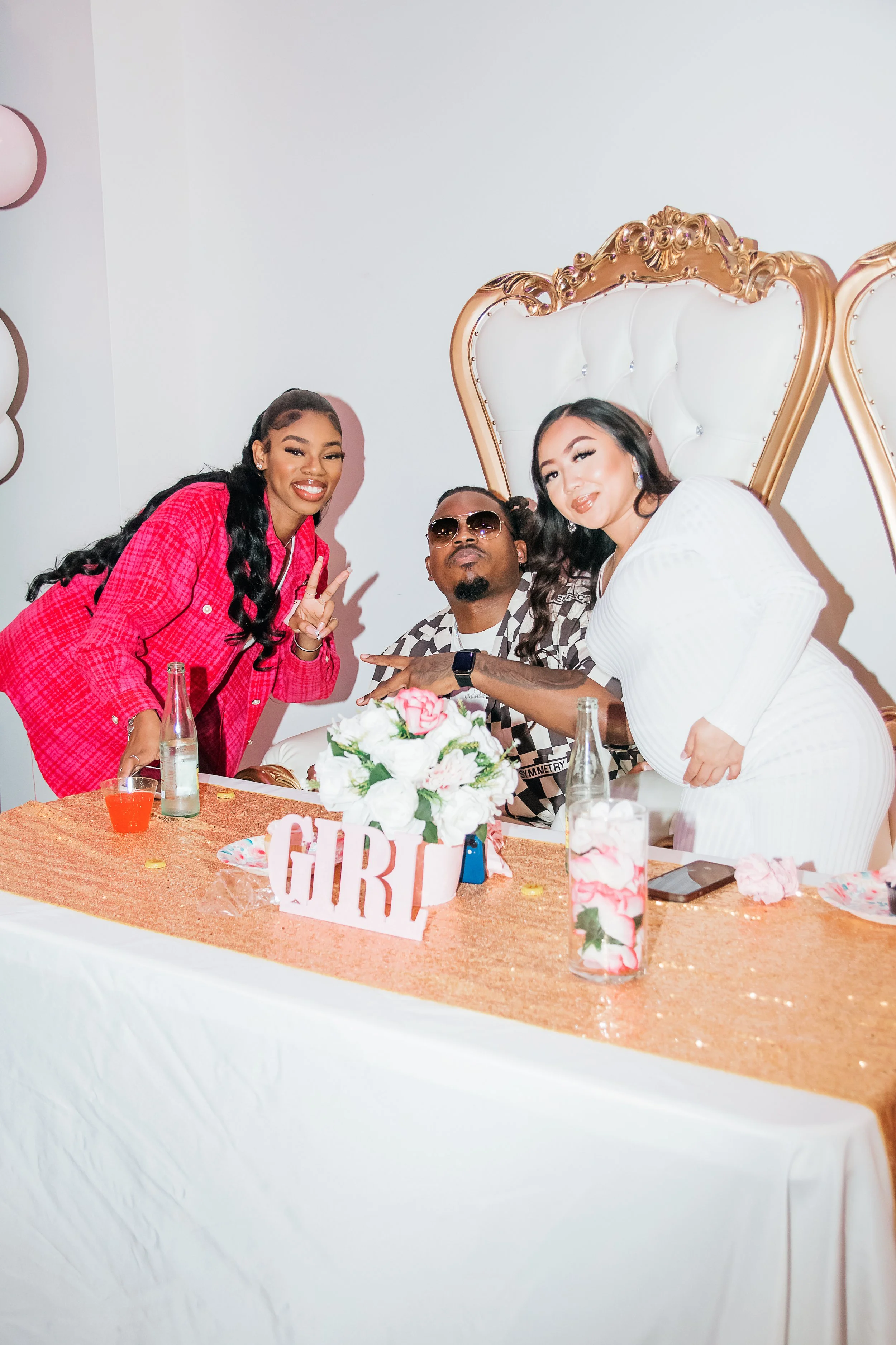Two women and a man seated at a decorated table with a large white and gold throne in the background, celebrating a girl-related event.