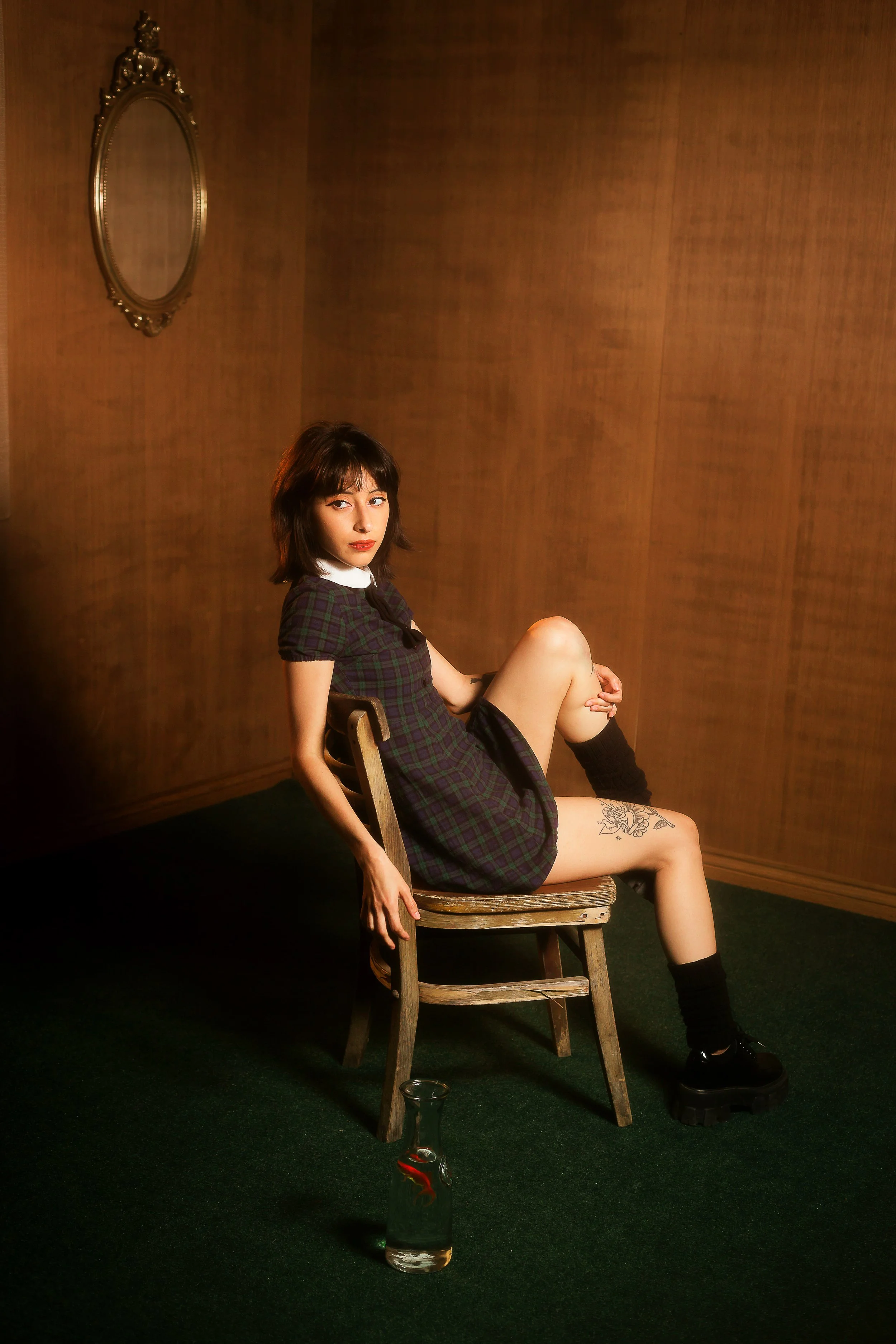 A young woman in a checkered dress with a white collar, sitting sideways on a wooden chair in a room with wooden walls and green carpet, next to a glass bottle with red liquid inside.