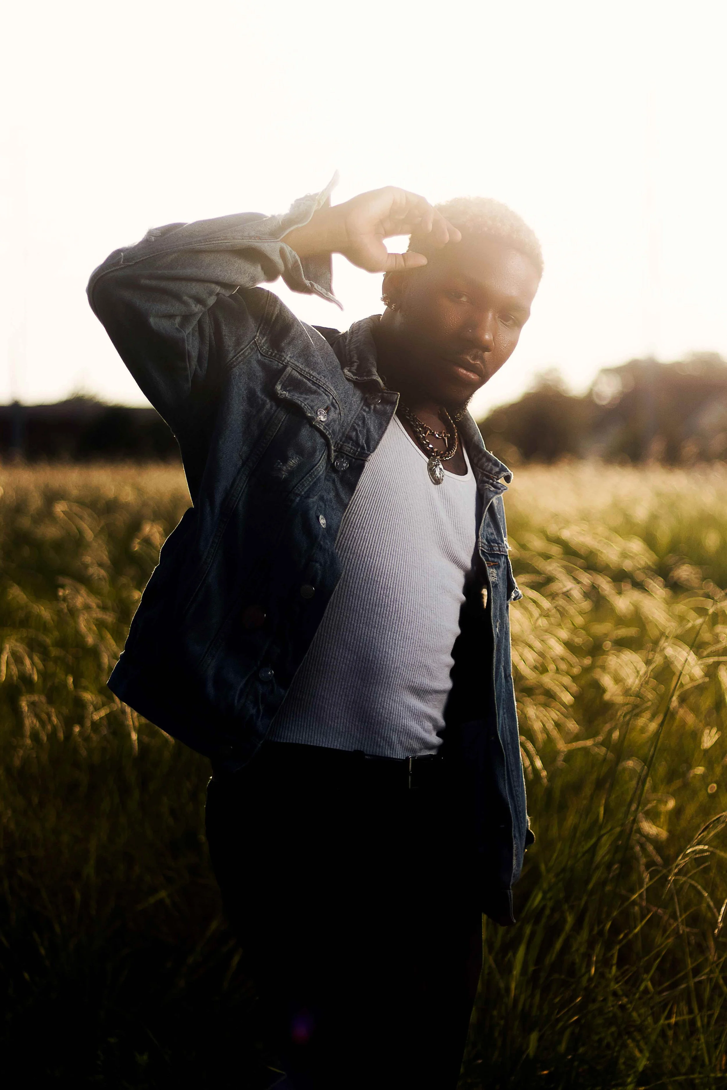 Sunset editorial portrait session in Houston, Texas by Lev’s Photography featuring a person wearing a denim jacket, white tank top, and layered necklaces standing in a field during golden hour