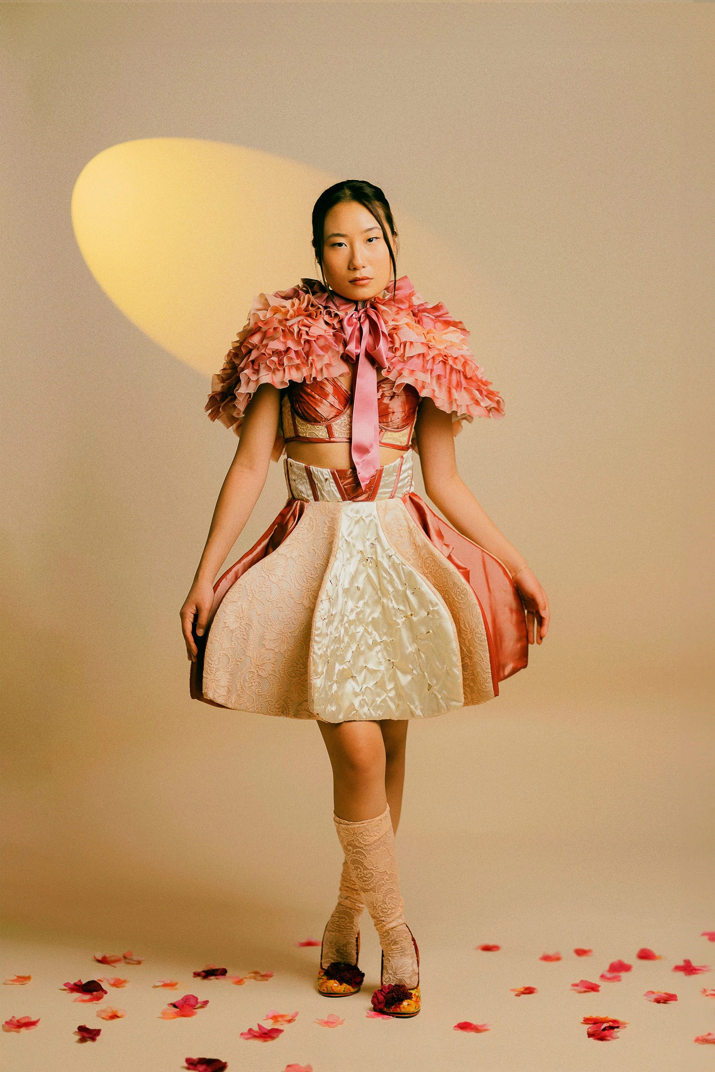 A woman standing in a beige studio with pink and red floral patterned high heels, lace knee-high socks, and a vibrant, layered, multicolored dress with ruffled pink shoulders and waist details. She has dark hair tied back and faces the camera with a
