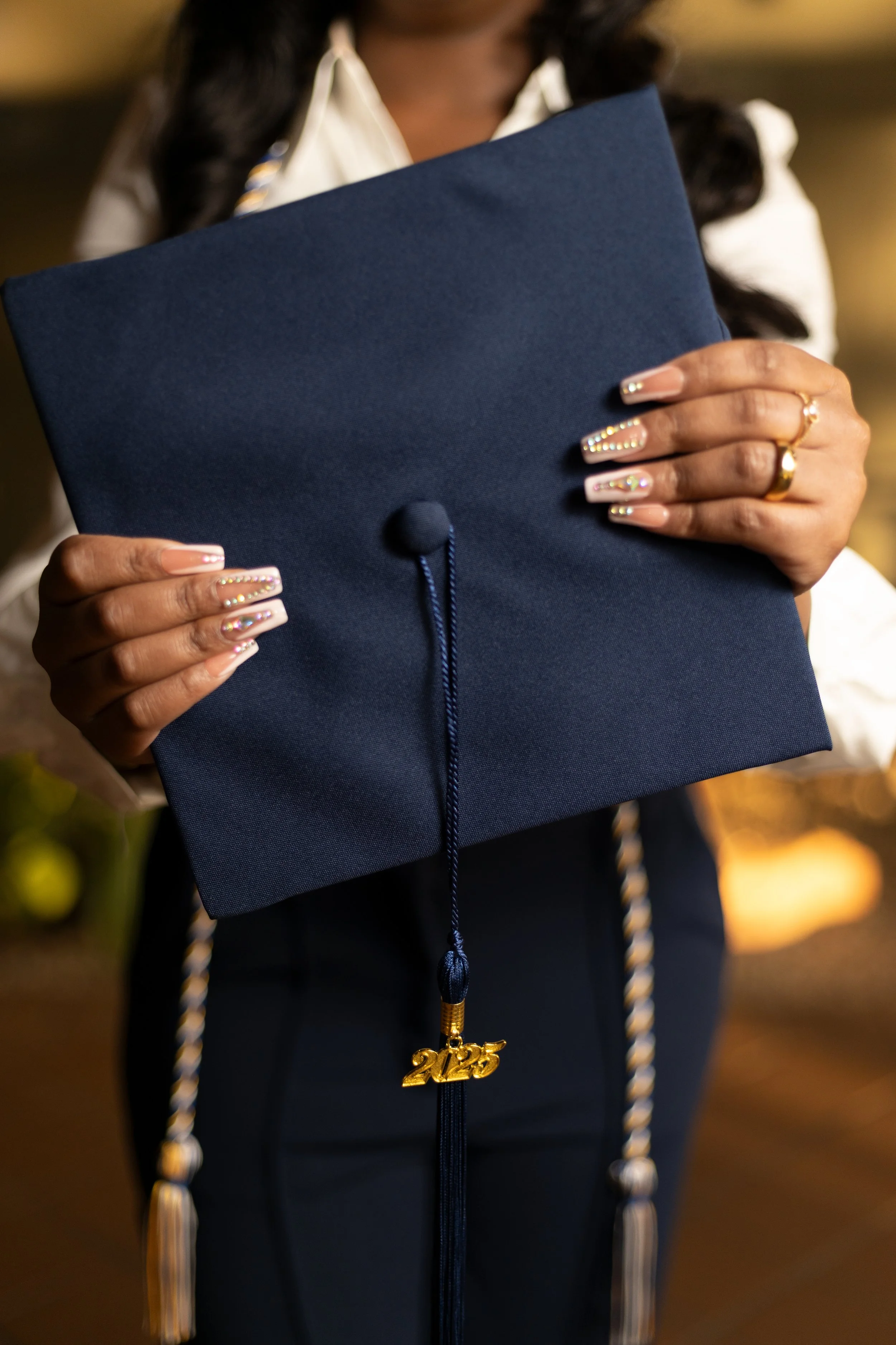 Person holding a navy graduation cap with a year charm hanging from the tassel, wearing rings and decorated nails, at a graduation ceremony.
