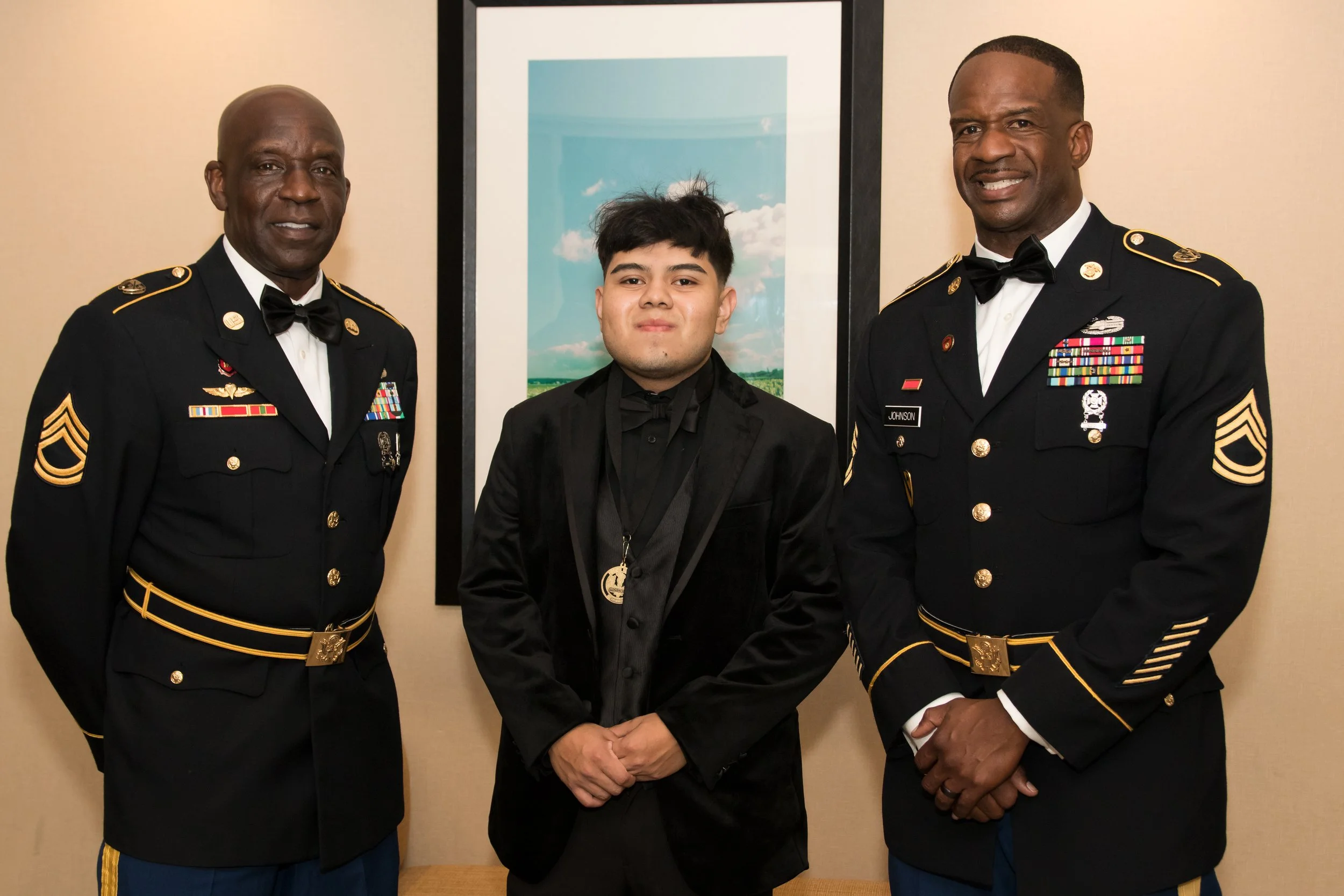 Three men in military dress uniforms and a young man in a black velvet tuxedo standing in front of a framed picture of a sky with clouds.