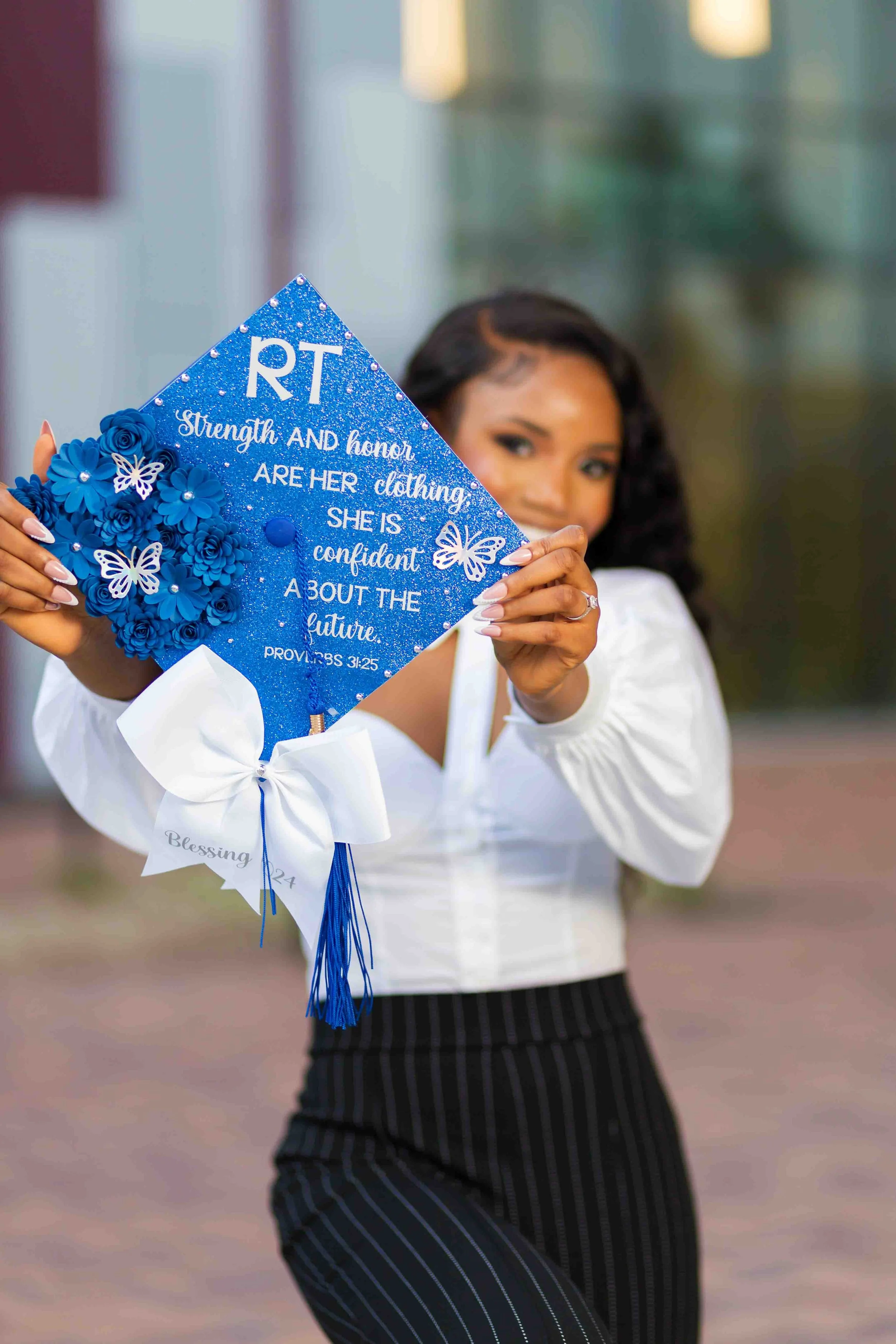 Outdoor graduation portrait of a woman holding a graduation cap decorated with blue flowers, butterflies, and a Bible verse, wearing a white blouse and black striped pants, smiling at the camera