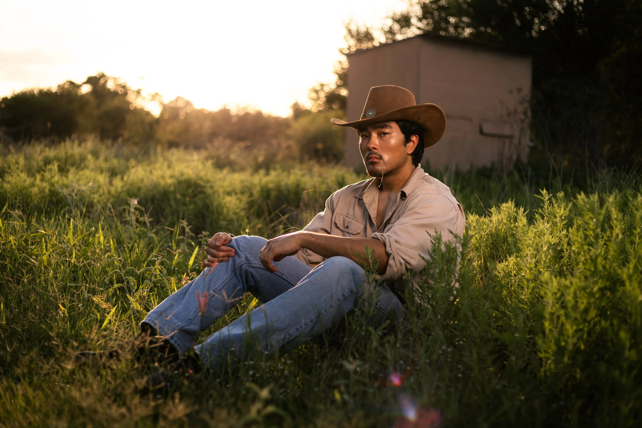 A man sitting in tall grass during sunset, wearing a cowboy hat, beige shirt, and jeans, with a serious expression.