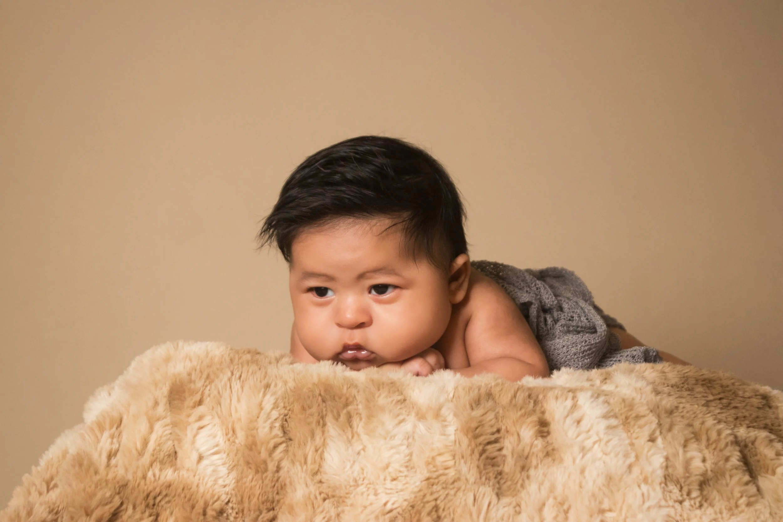 A young child with dark hair lying on a plush, tan blanket with their chin resting on their folded arms, looking to the side with a serious expression.