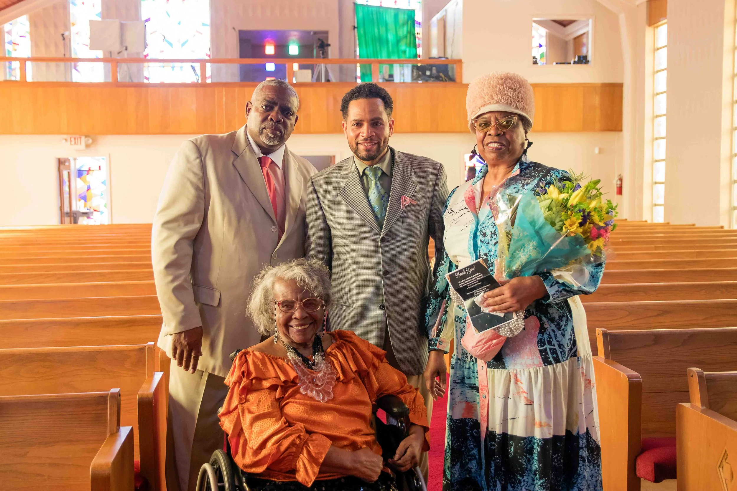 Five people, including an elderly woman in a wheelchair, standing inside a church with wooden pews. The elderly woman is smiling, wearing an orange dress, glasses, and a beaded necklace. The woman on the right is holding a bouquet of flowers and a th