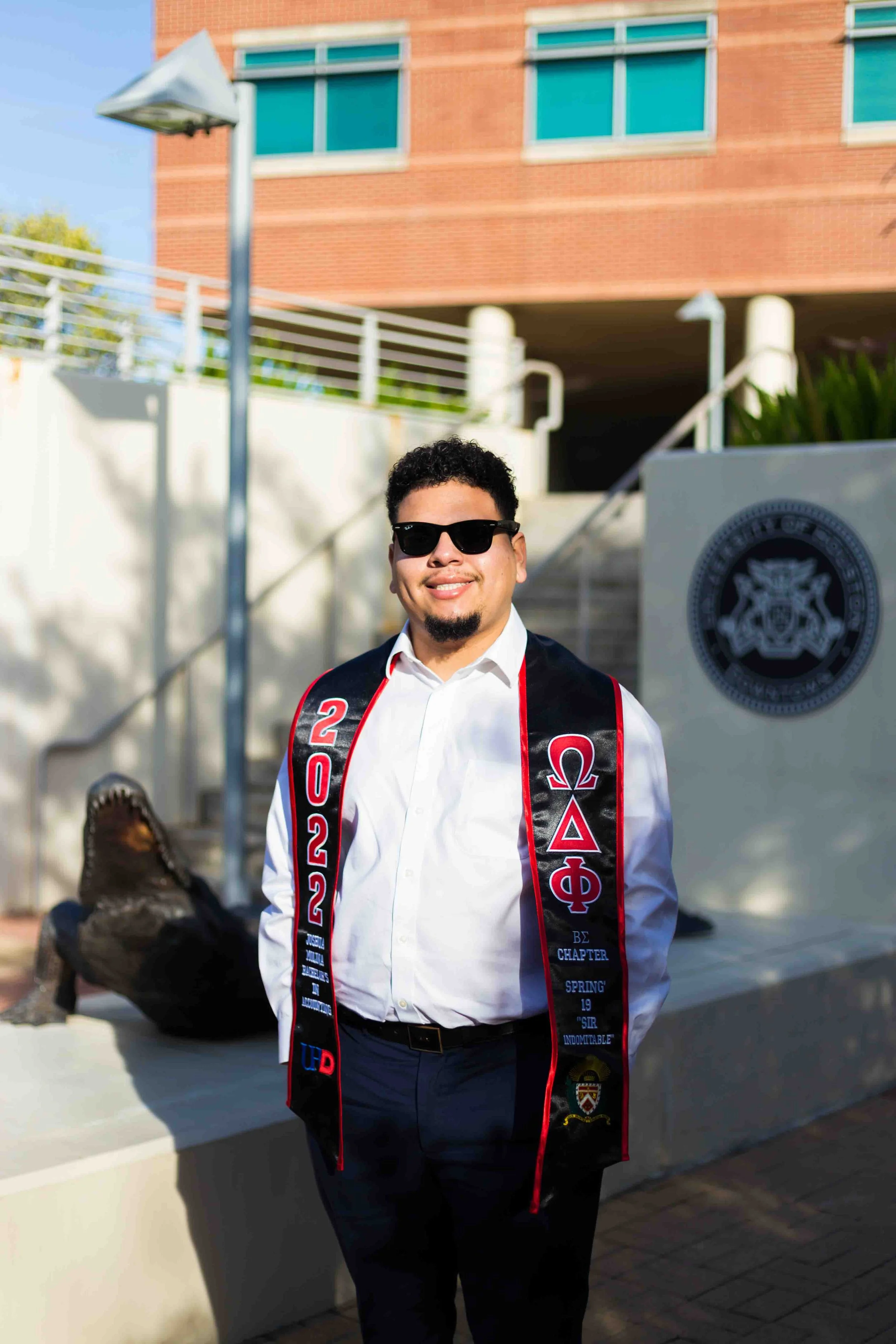 University of Houston–Downtown Marilyn Davies College of Business graduate portrait featuring a student in cap and gown standing outdoors on campus, celebrating their business school graduation