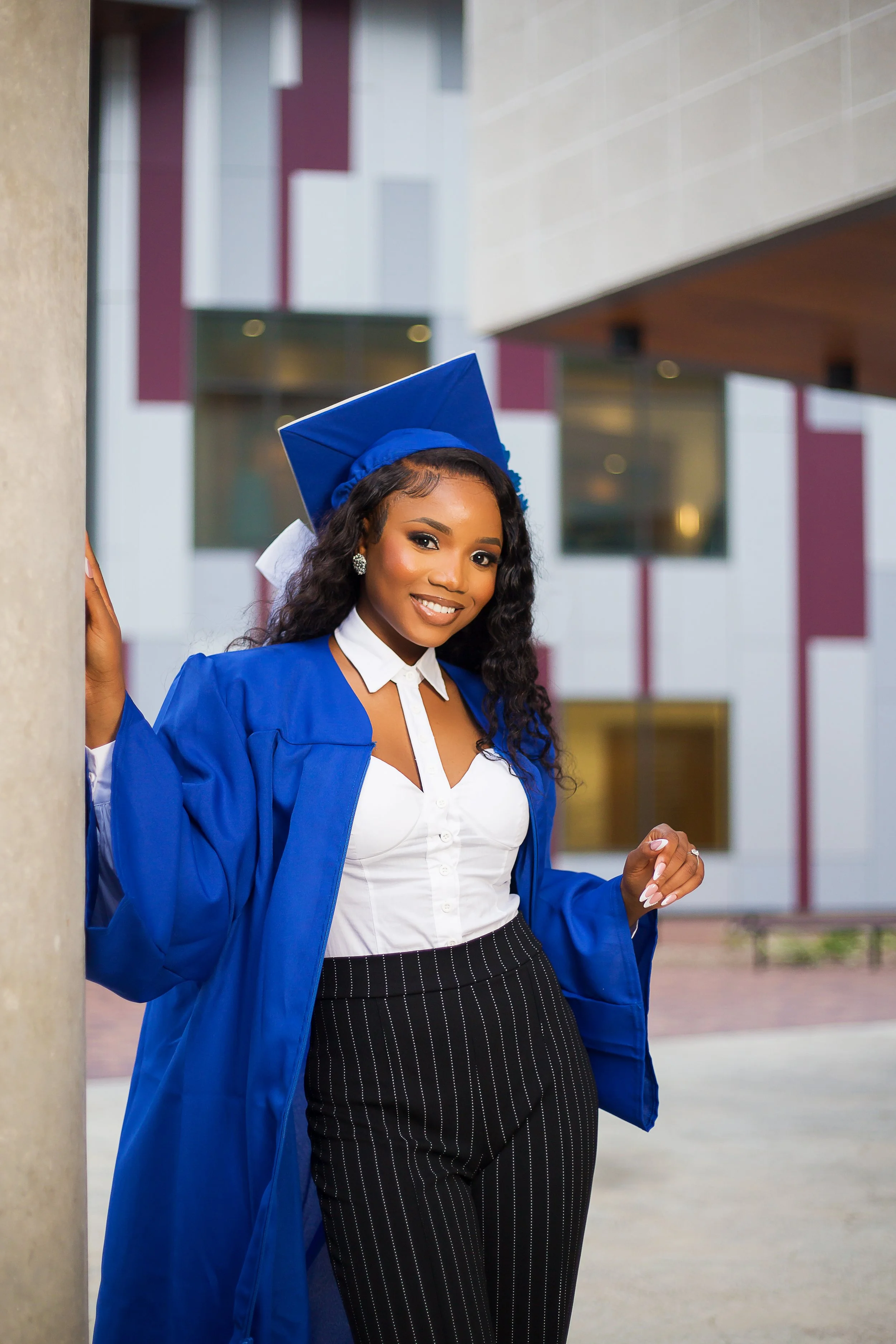 A young woman in a blue graduation gown and cap, smiling and posing outdoors in front of a modern building.