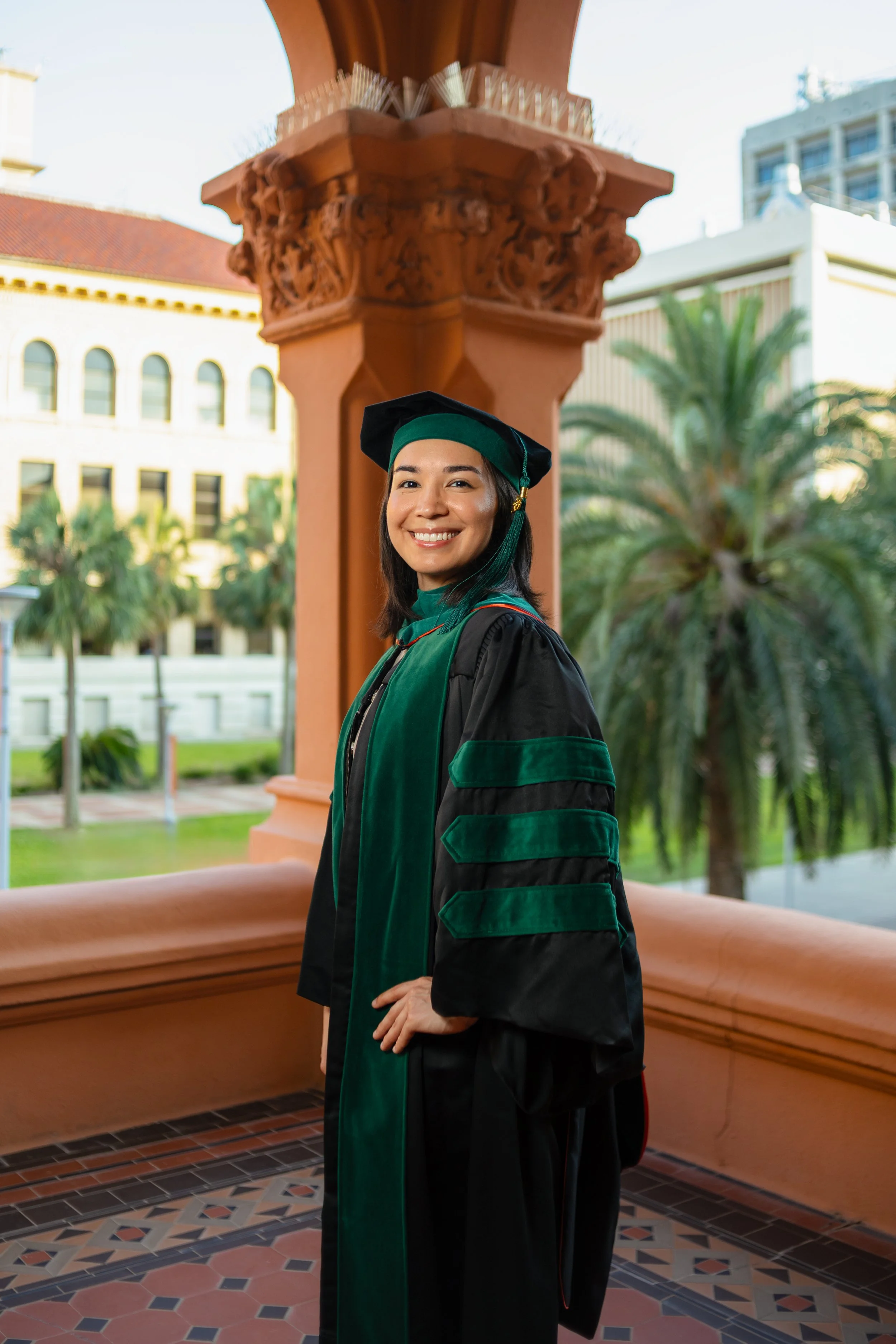 A young woman in academic regalia standing outdoors, smiling, with palm trees and a building in the background.