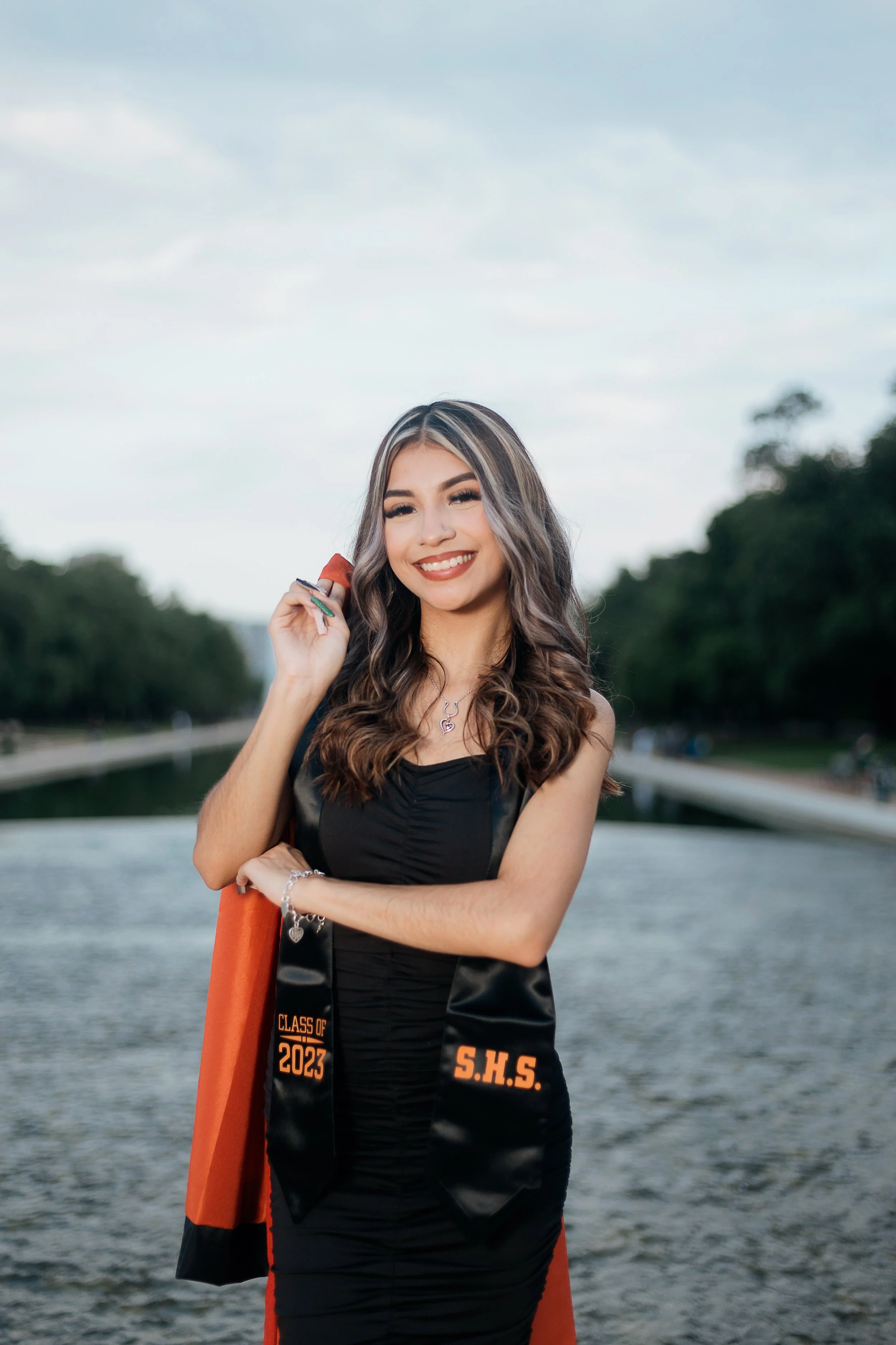A young woman in a graduation gown celebrating outdoors near a river with trees in the background, smiling, holding a diploma, and wearing a necklace and bracelet.