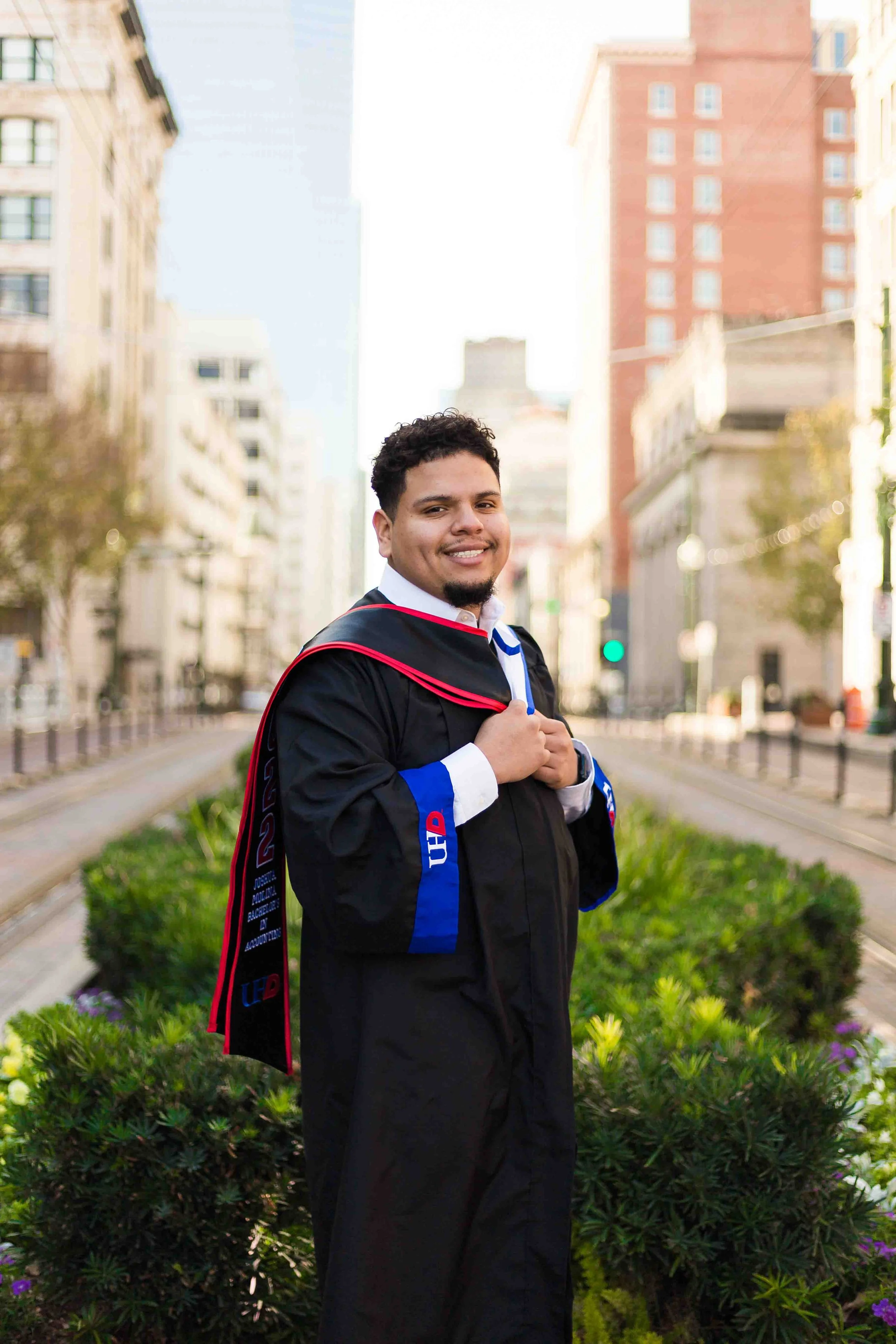 Graduation portrait at University of Houston–Downtown featuring a smiling young man in a cap and gown standing outdoors in an urban setting with downtown Houston buildings in the background