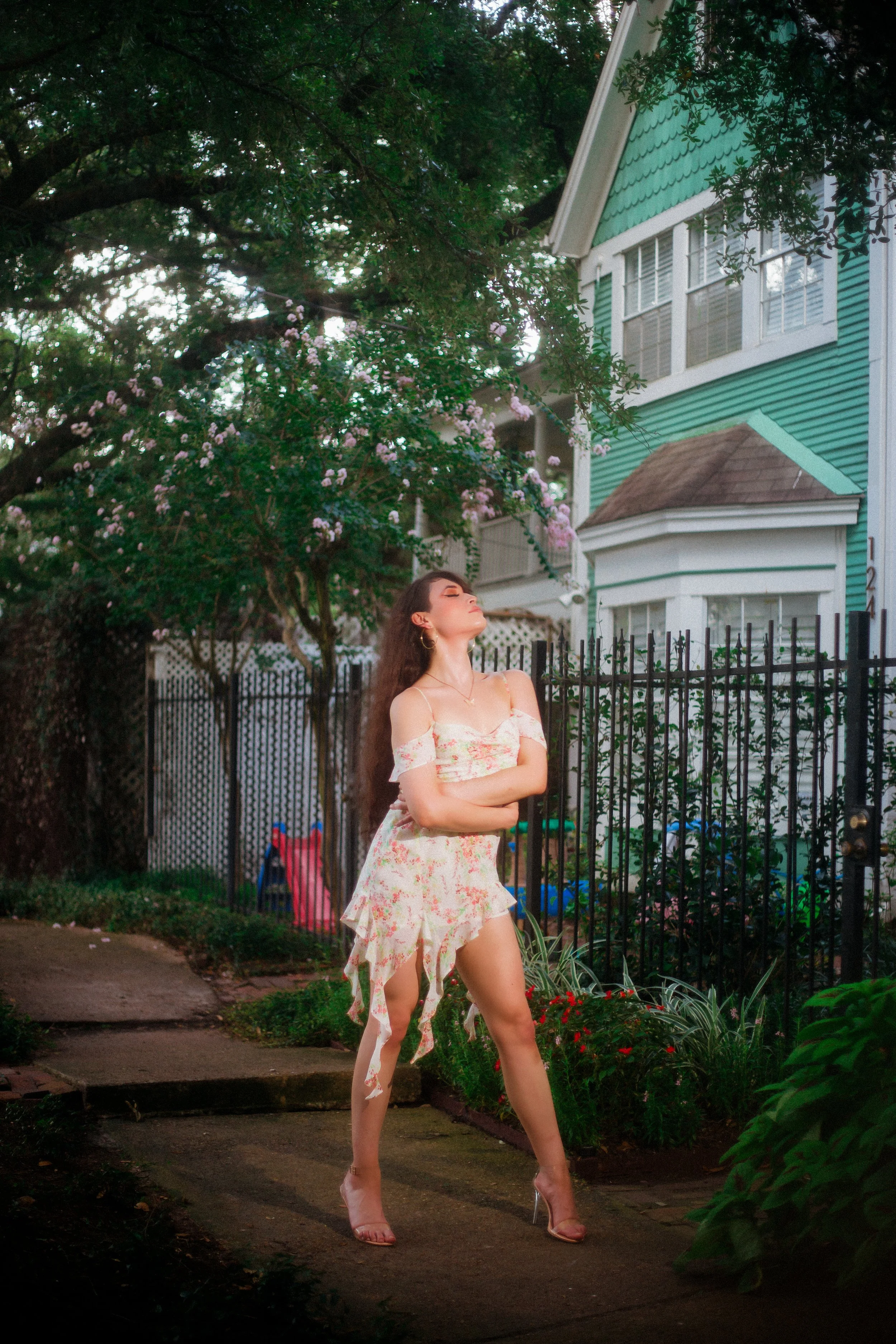 A woman in a floral off-the-shoulder dress and clear high heels posing with arms crossed on a garden path in front of a black metal fence and a turquoise house, with flowering trees in the background.
