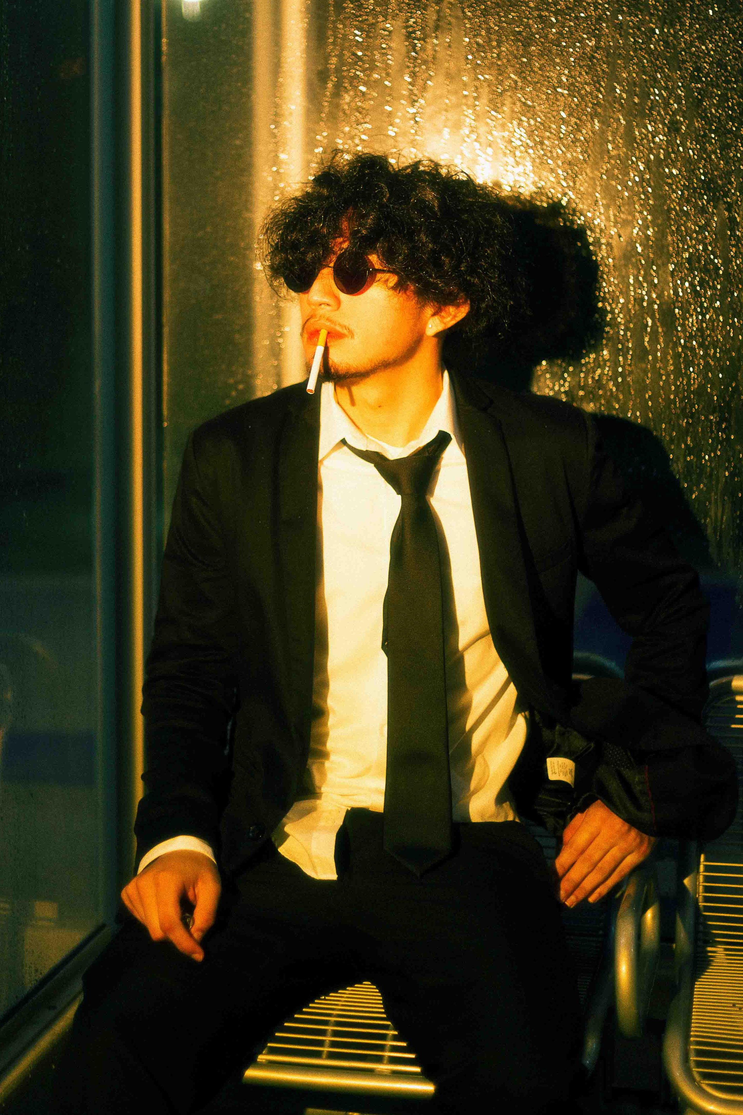 Houston cinematic portrait of a man with curly black hair, sunglasses, and a cigarette in his mouth, wearing a black suit and tie, sitting on a bench at night with a metallic rainy background illuminated by warm yellow light