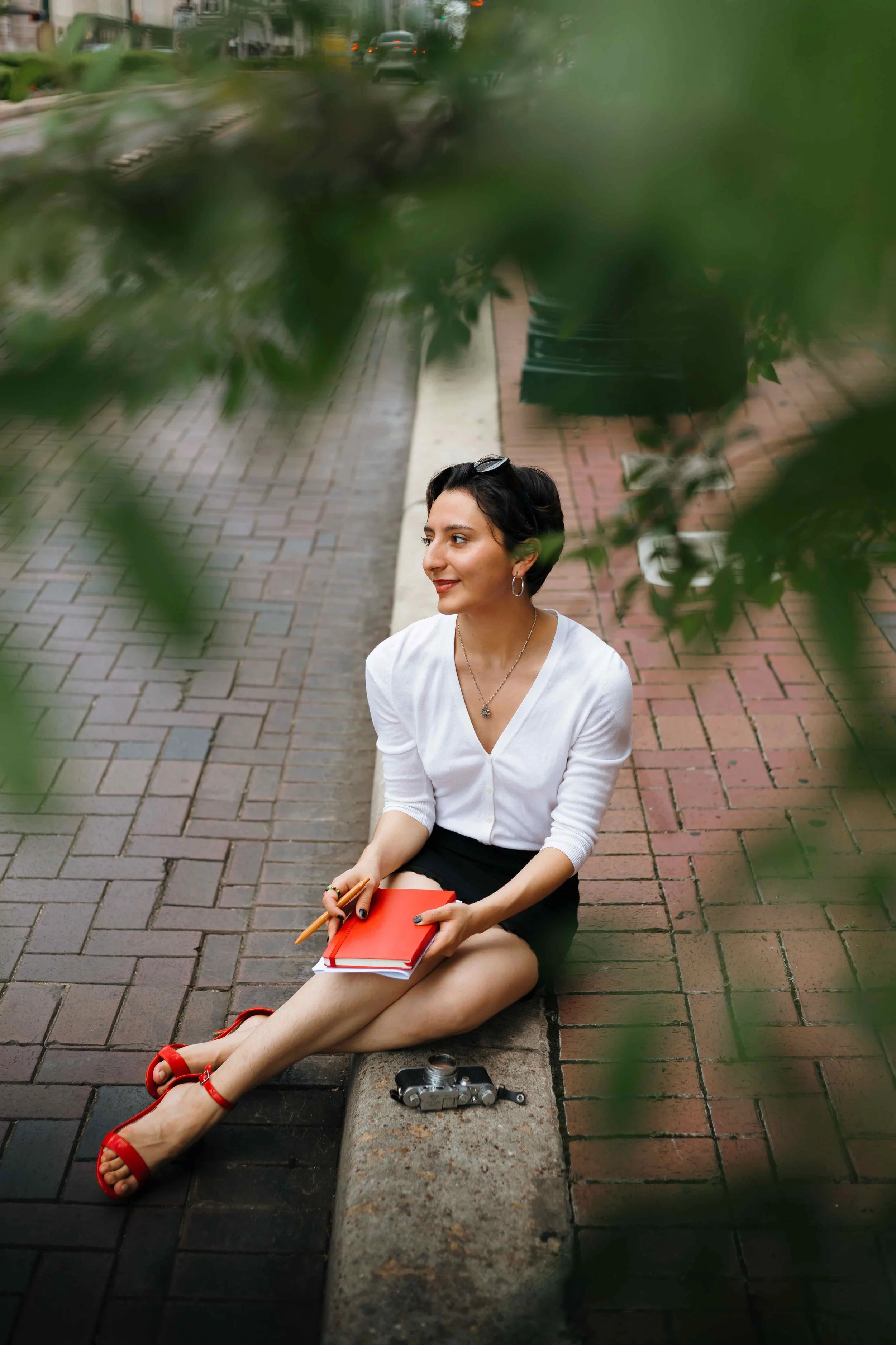 Houston branding photography session by Lev’s Photography featuring a woman with short black hair sitting outside a coffee shop with a vintage camera and red notebook on a brick sidewalk framed by green leaves.