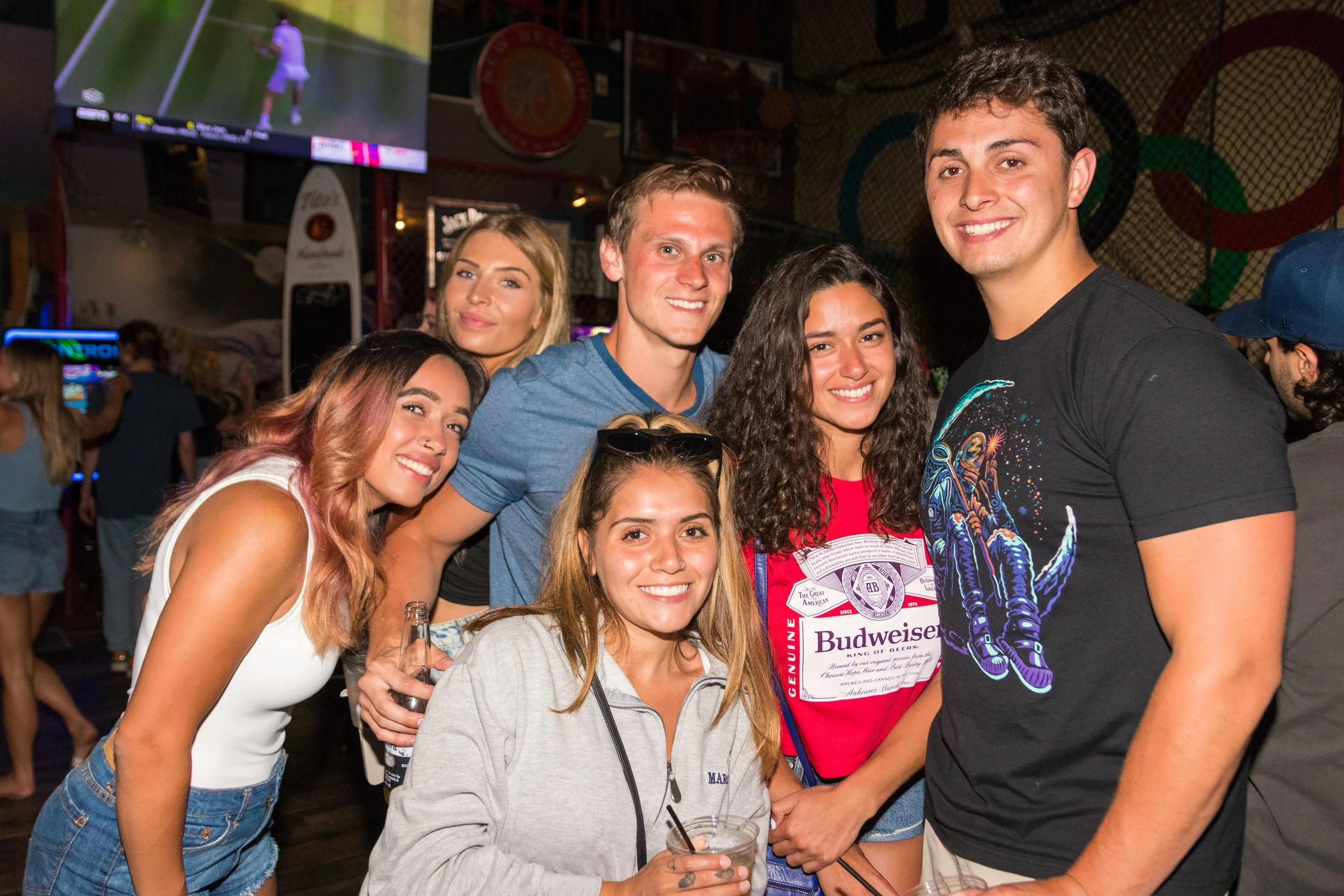 Group of six young friends with smiling faces enjoying a night out at a bar or club, some holding drinks, in a lively atmosphere with a TV screen and neon lights in the background.