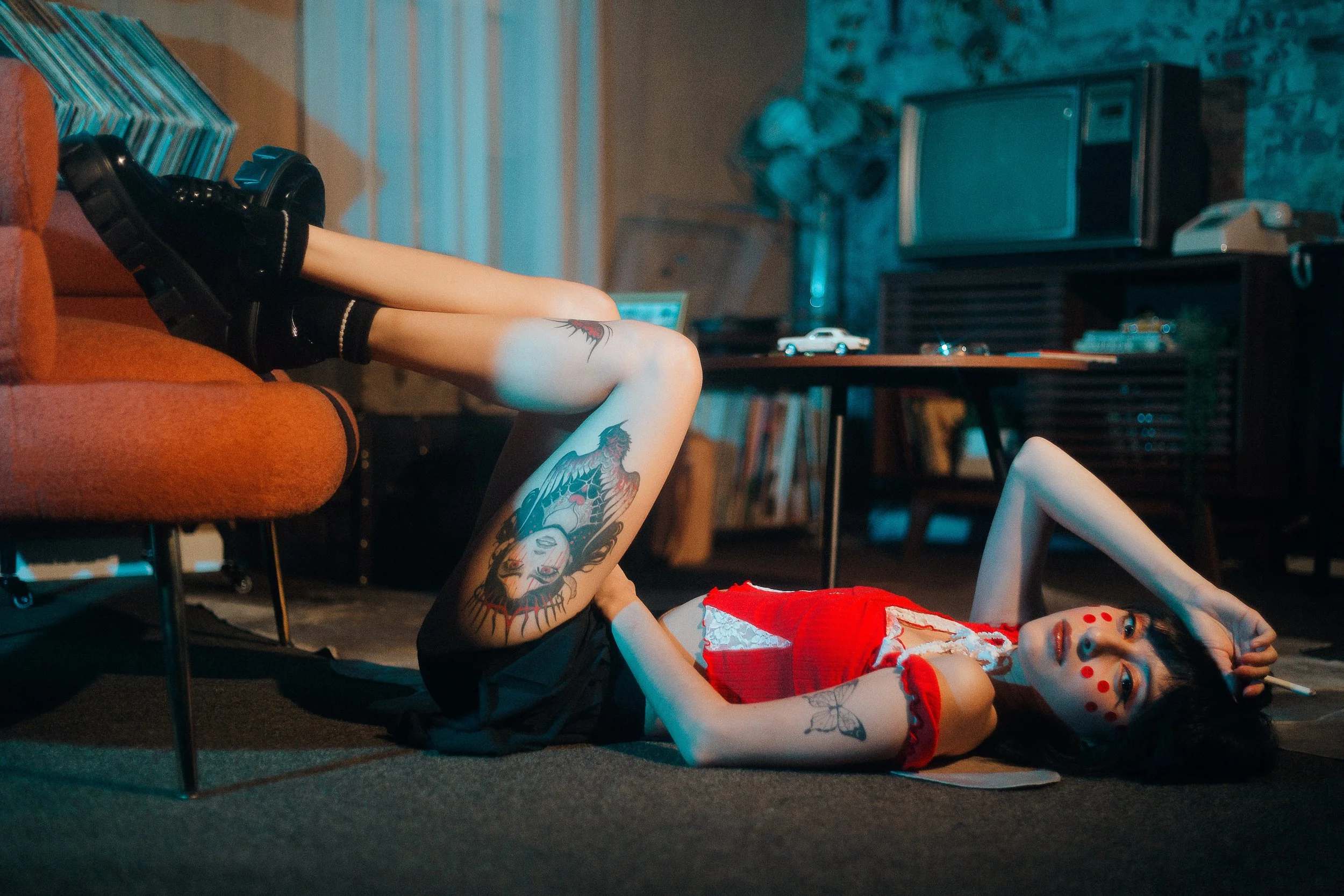 A woman with tattoos and face paint lying on the floor of a room with vintage decor, wearing a red top, black skirt, and black boots, with her legs resting on an orange sofa.