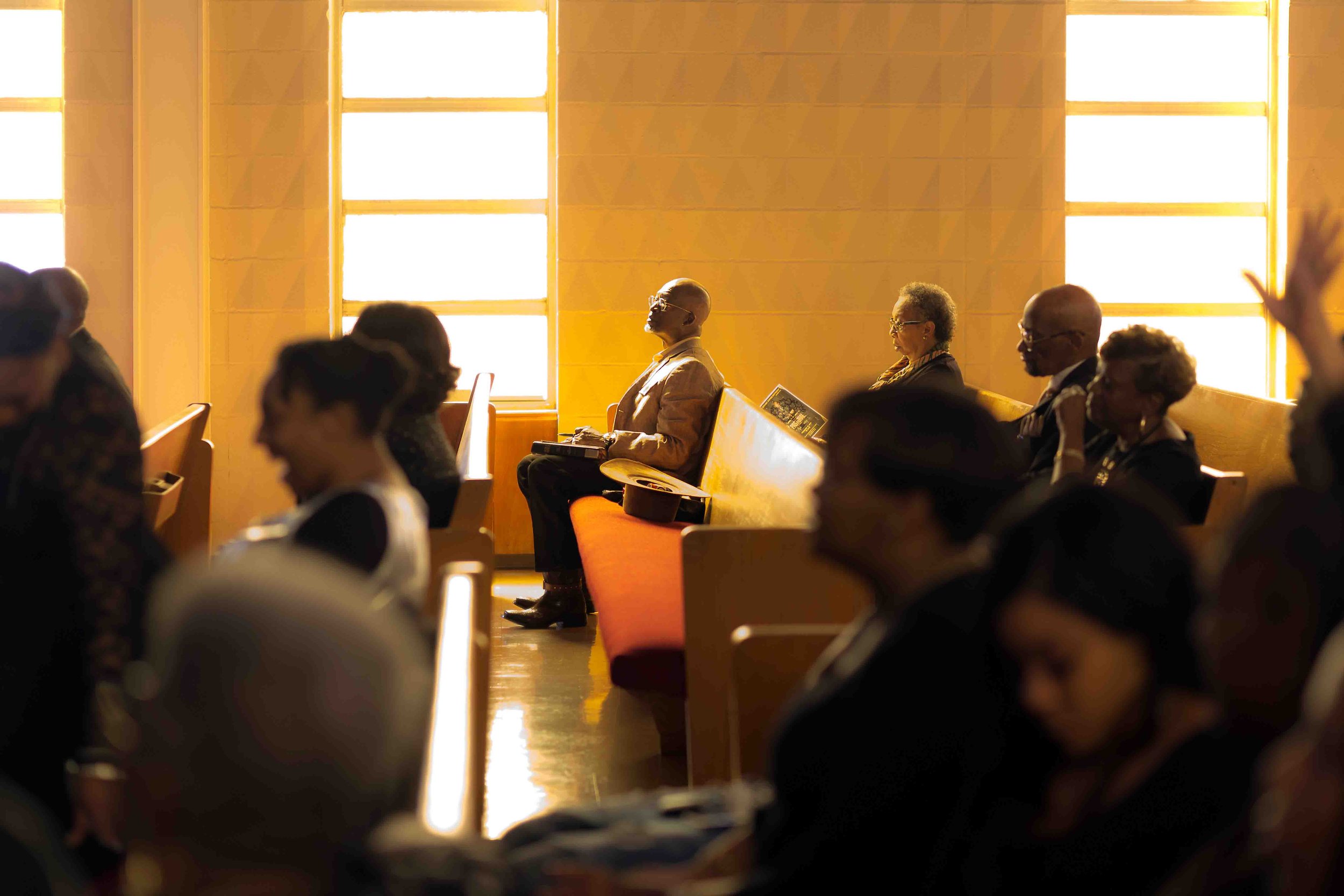 Houston church service documentary by Lev's Photography featuring congregants seated in wooden pews with warm sunlight streaming through large stained-glass windows, capturing candid moments of worship, community, and spiritual reflection during Sund