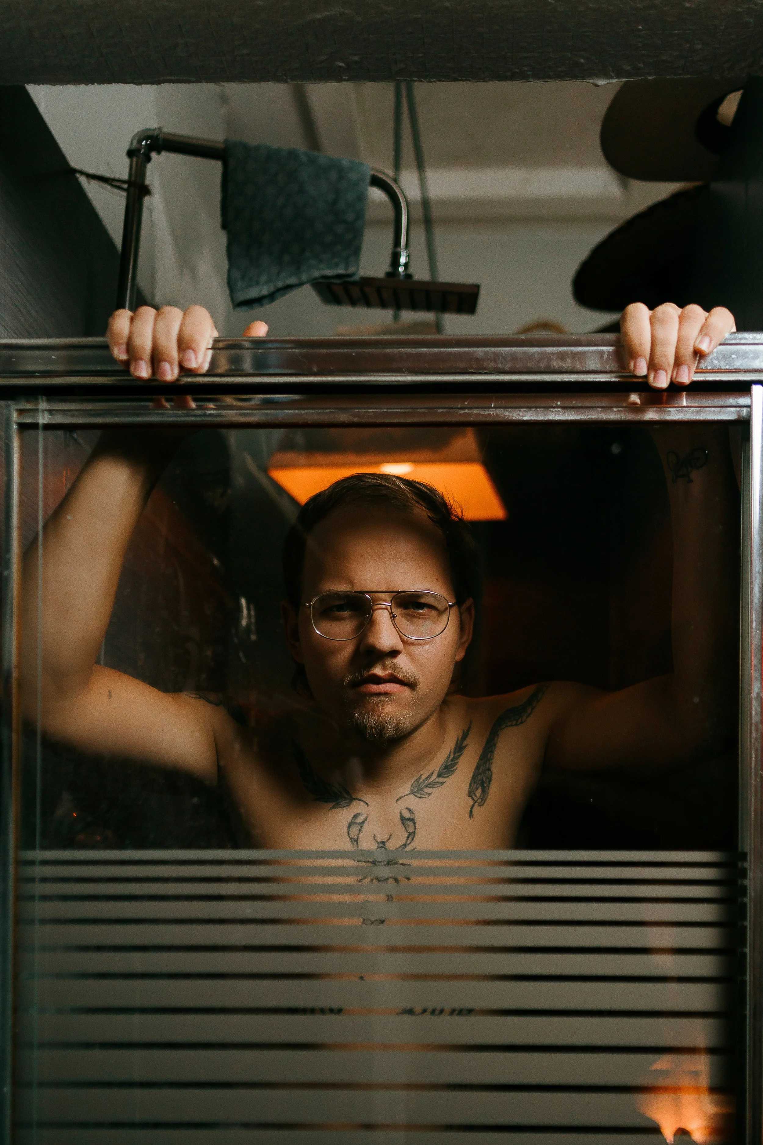 A shirtless man with glasses and tattoos looking through a glass display case, with his hands gripping the top of the case and a shower in the background.