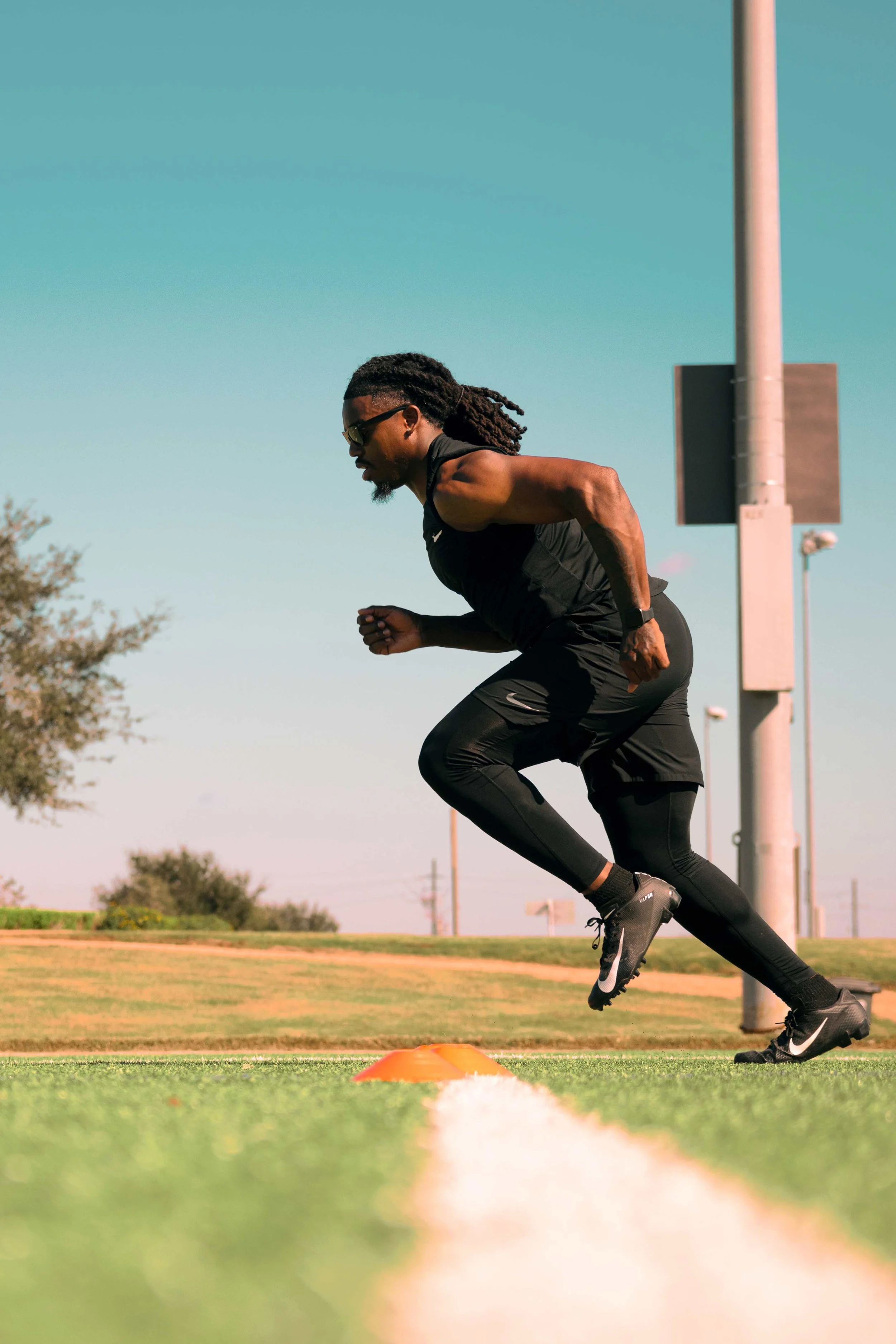 Athletic sports portrait in Houston, Texas by Lev’s Photography featuring a male athlete in black running gear and sunglasses sprinting on an outdoor track with an orange cone and cloudy sky, ideal for football, NFL, or fighter branding.
