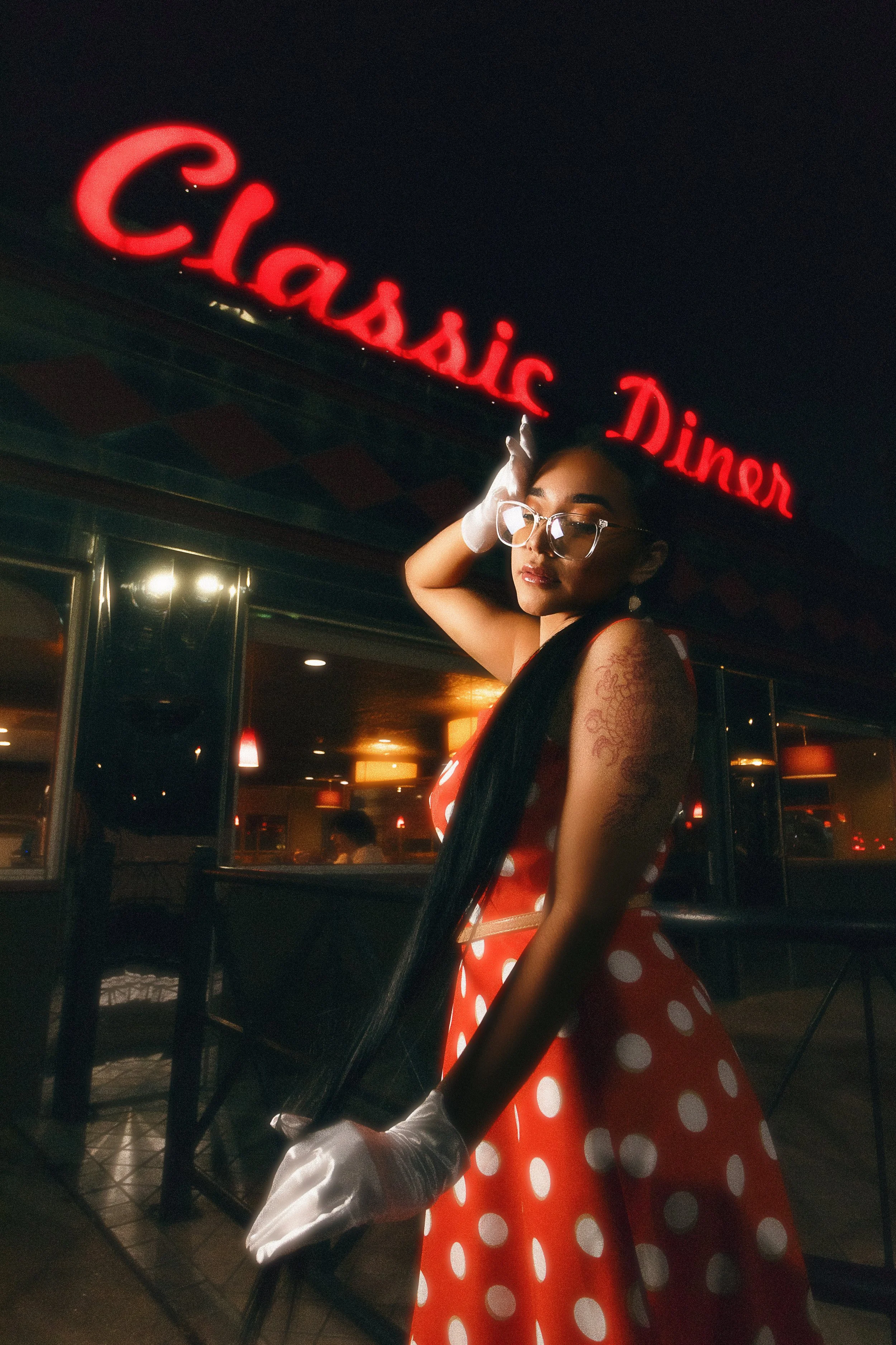 A woman with long black hair, glasses, and tattoos on her arm, dressed in a red polka dot dress with white gloves, posing inside a vintage diner with a neon sign above that reads 'Classic Diner' at night.