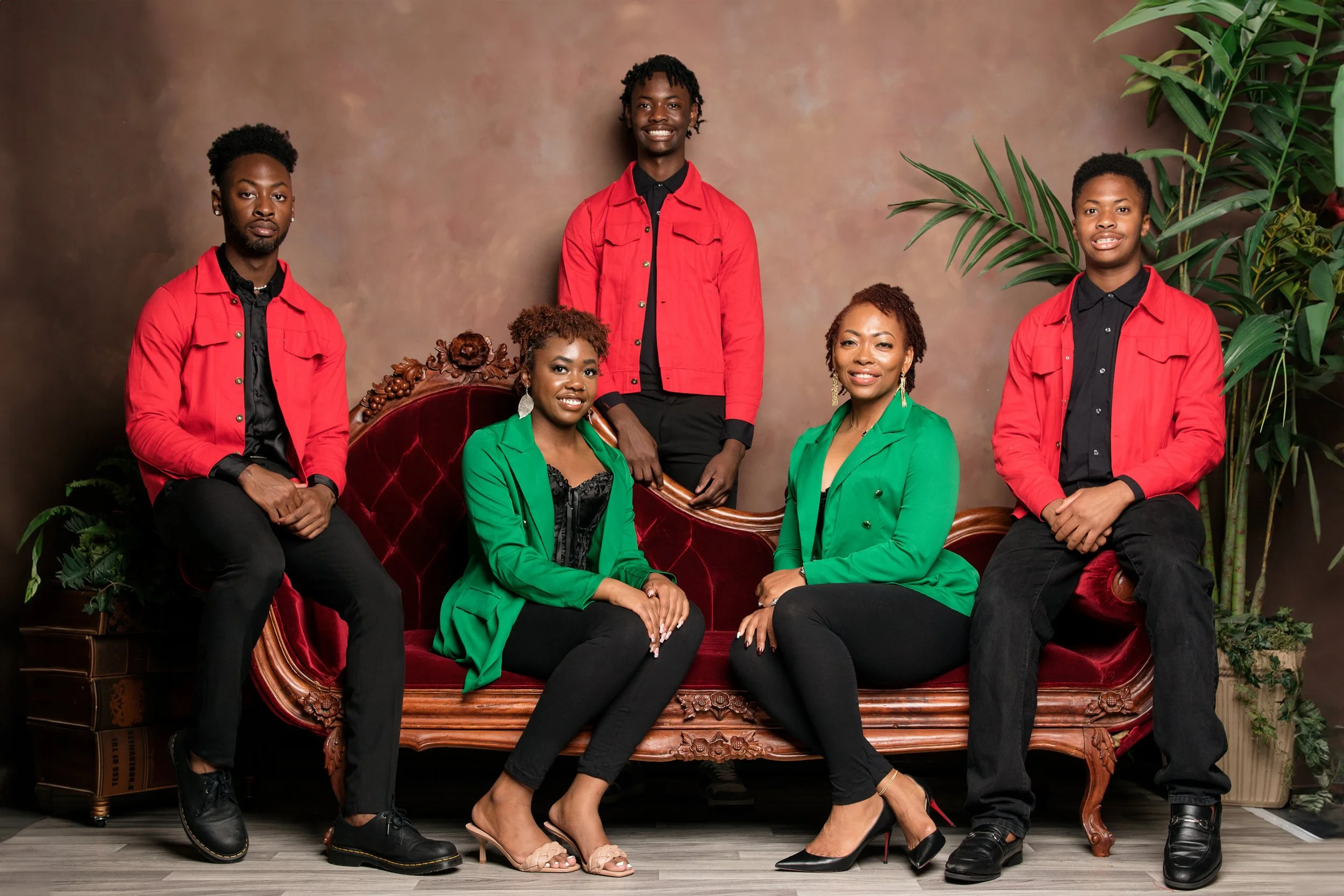 Group of five people, three women and two men, posing in a studio with a vintage sofa, dressed in matching red and green jackets and black pants, with a brown backdrop and green plants.