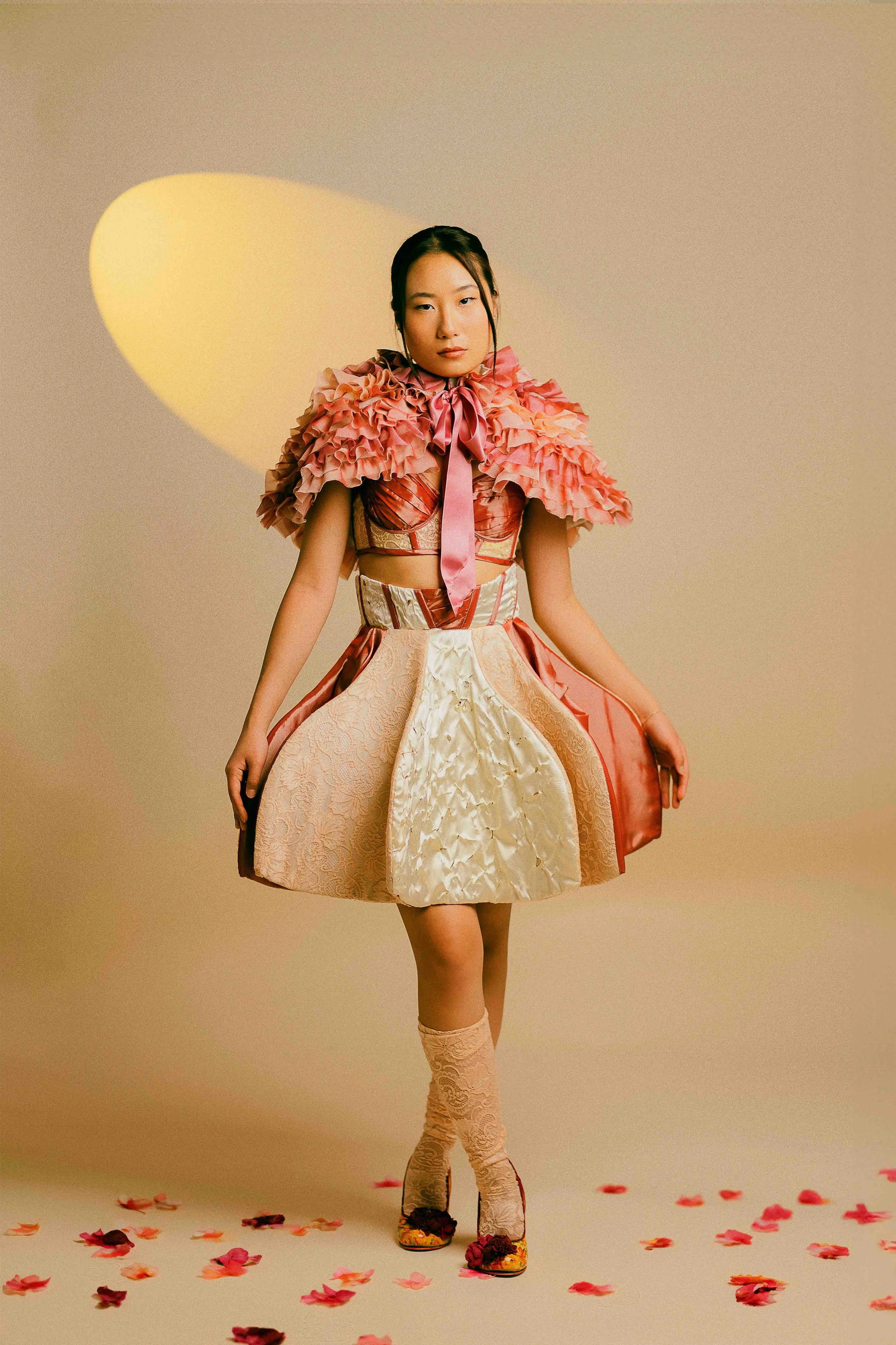 Editorial studio portrait of a young woman posing in a colorful, elaborate dress with ruffles and floral patterns, standing against a beige backdrop with scattered flower petals