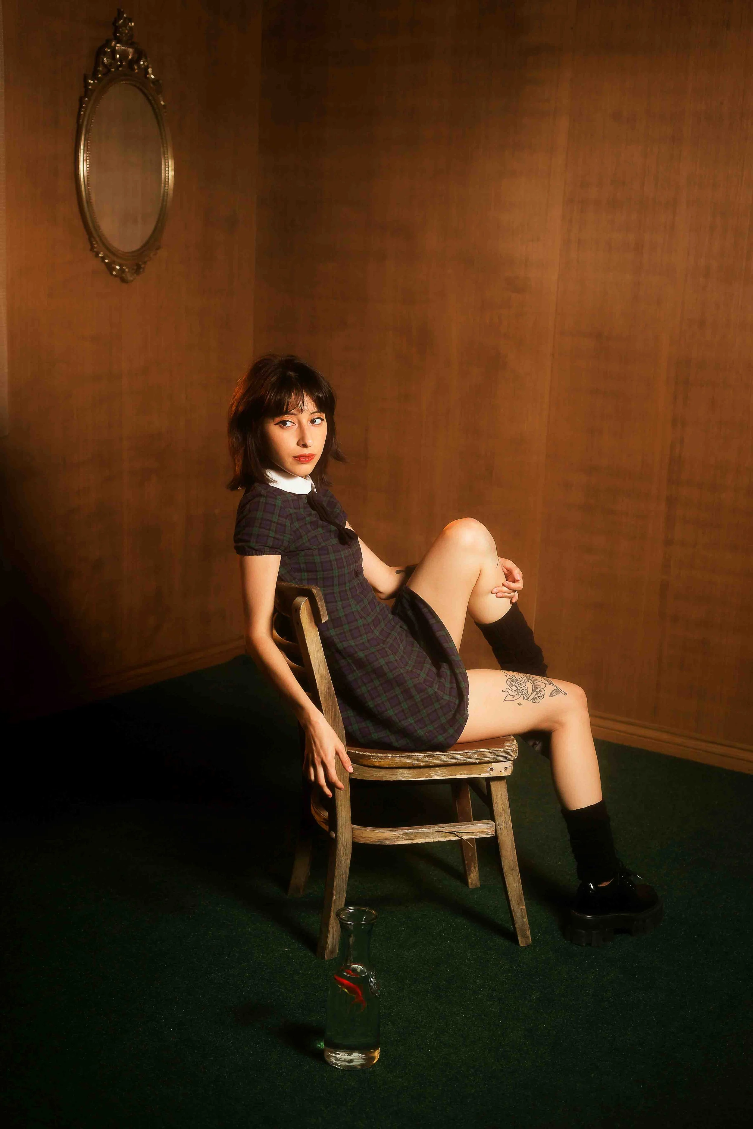 Houston editorial portrait of a young woman with short dark hair and a thigh tattoo sitting on a wooden chair in a room with brown wooden walls and dark green carpet, wearing a plaid dress, black socks, and black platform shoes, with a glass bottle c