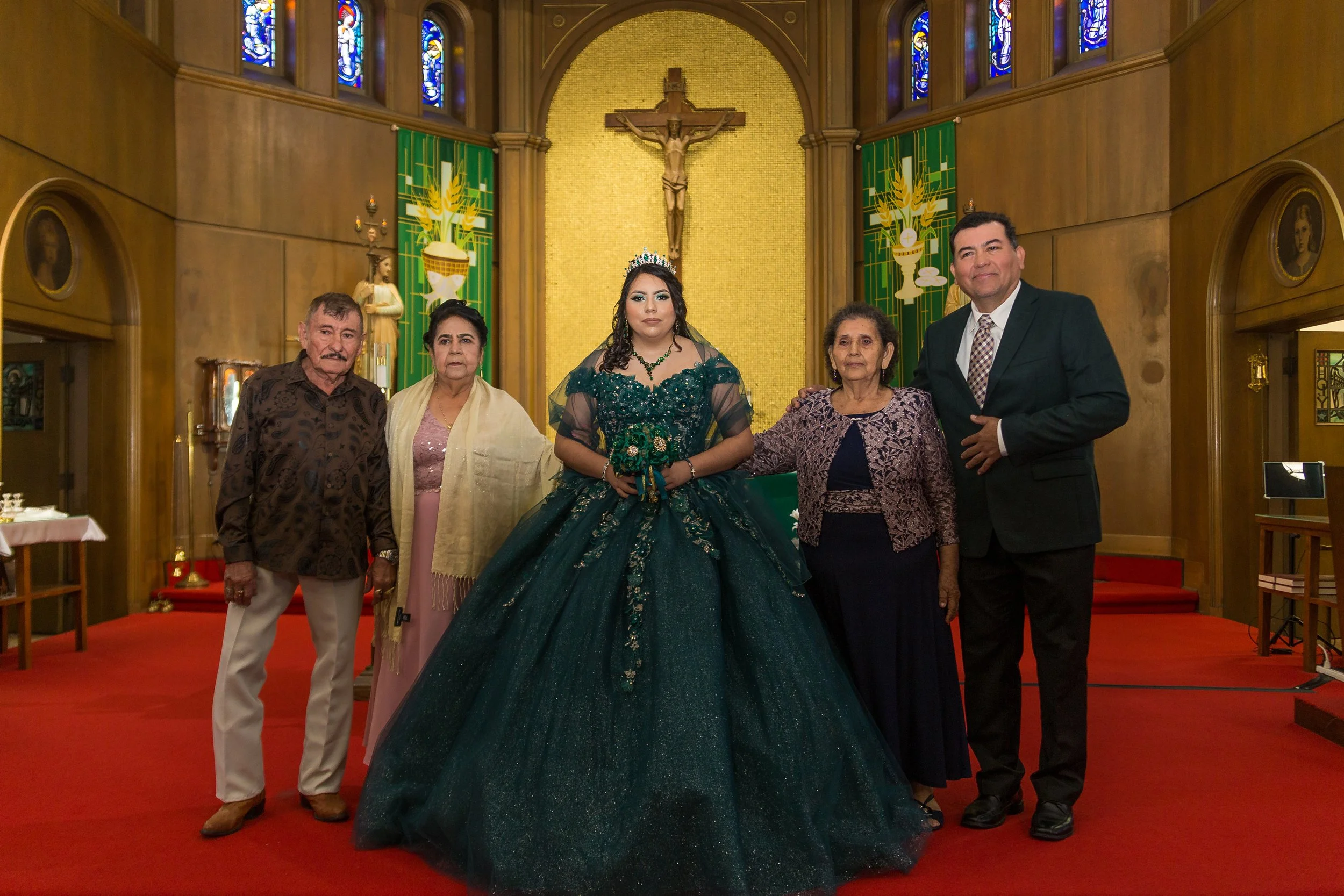 A large family poses for a photo inside a church. The young woman in the center wears a dark green ball gown and a tiara, indicating she's celebrating a special event like her quinceañera or prom. She is surrounded by four older adults, two women and