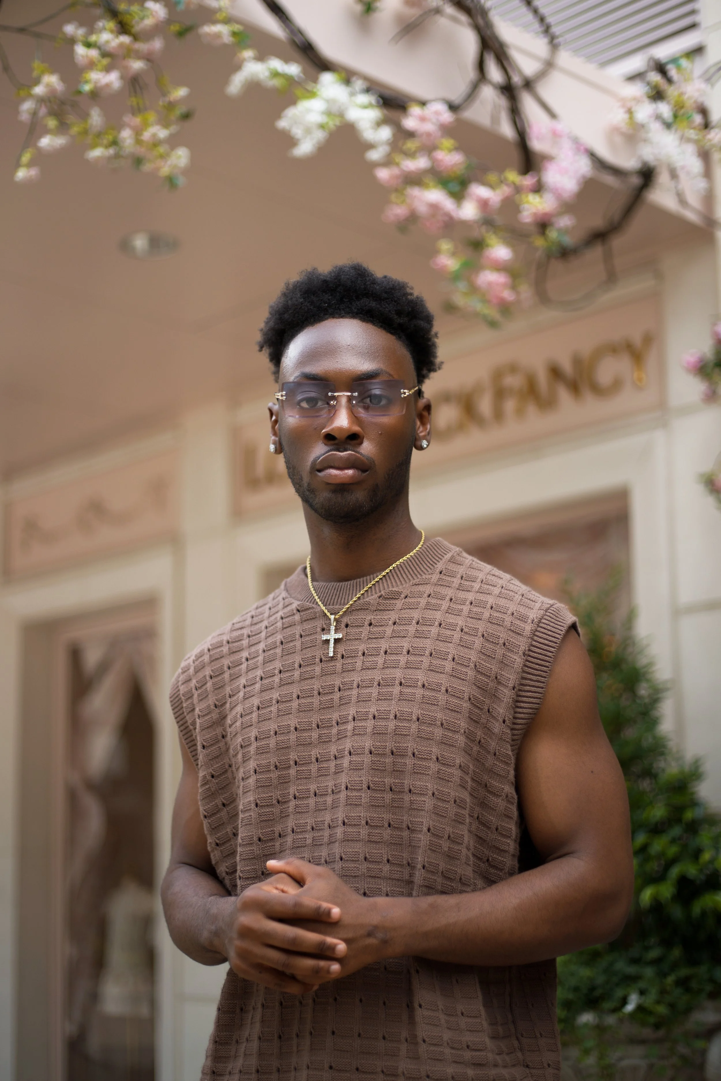 A young man with glasses, earrings, and a gold cross necklace standing outdoors in front of a building decorated with pink and white flowers.