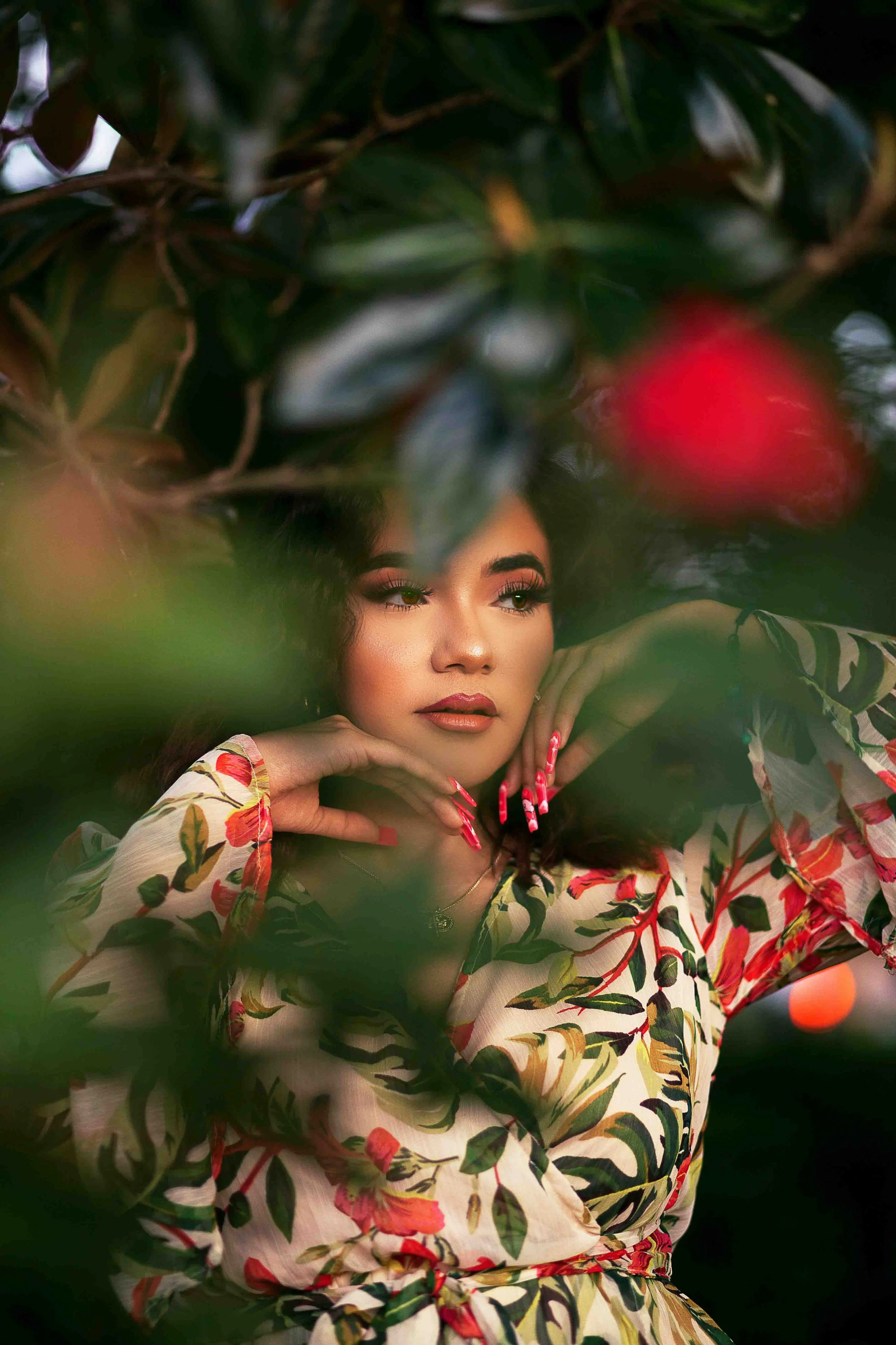 A woman with long dark hair and makeup, wearing a floral patterned dress, poses with her hands near her face among dense green leaves. Red and pink flowers are also visible in the background.