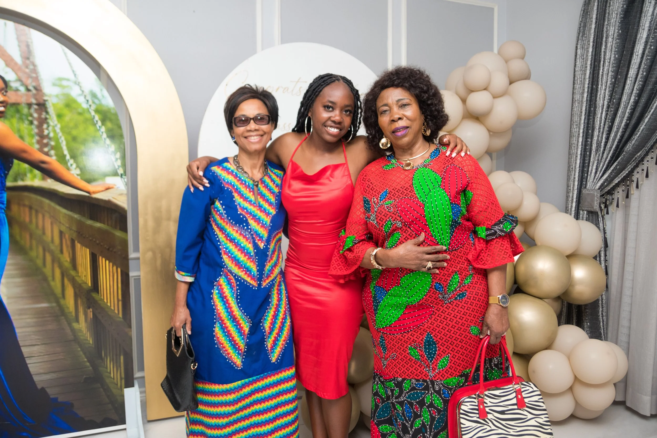 Three women standing together at a celebration, wearing colorful dresses, with balloons in the background.