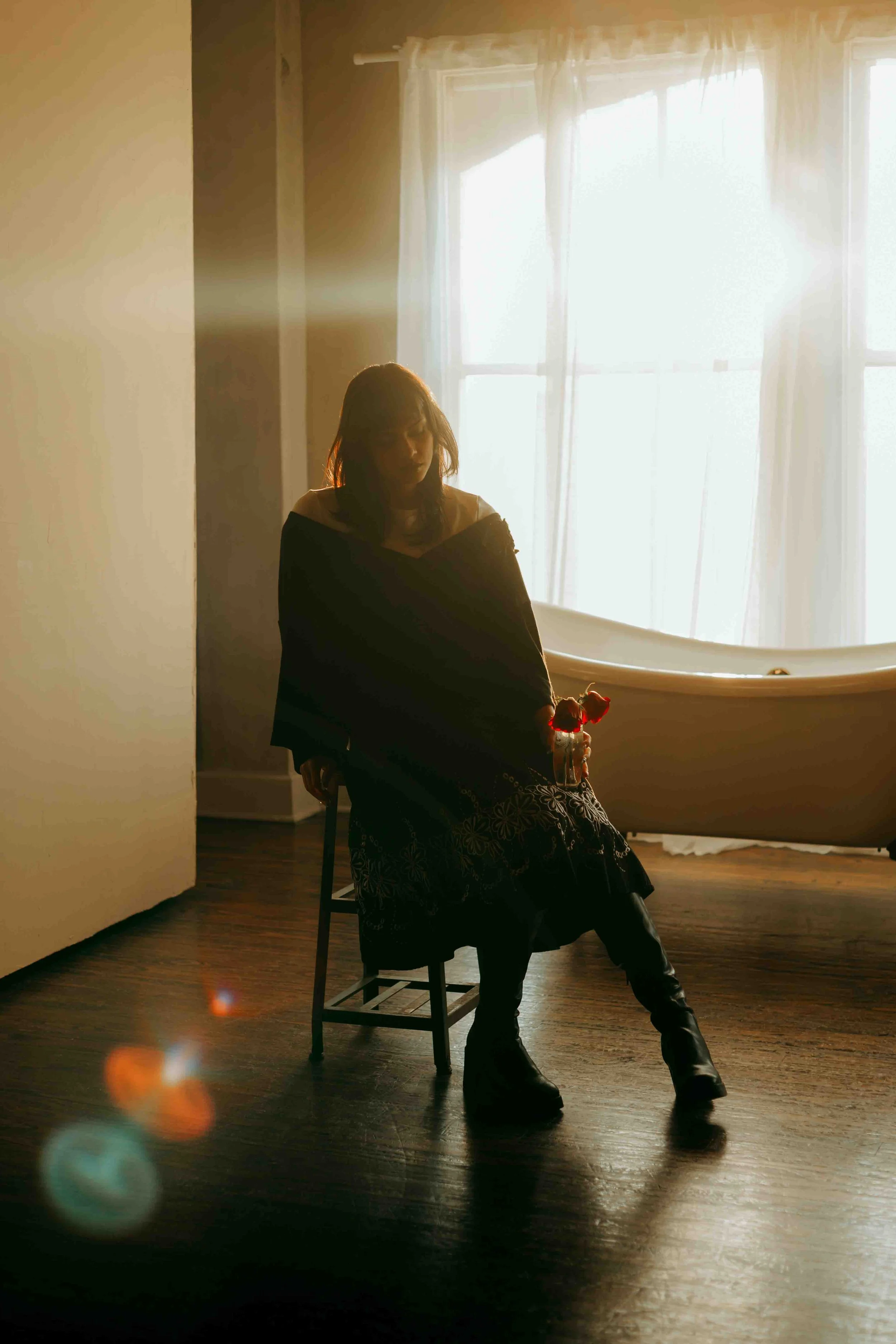 Backlit editorial portrait of a woman sitting on a chair in a sunlit room with wooden floors, holding a glass with flowers, wearing dark clothing and boots, with a bathtub in the background