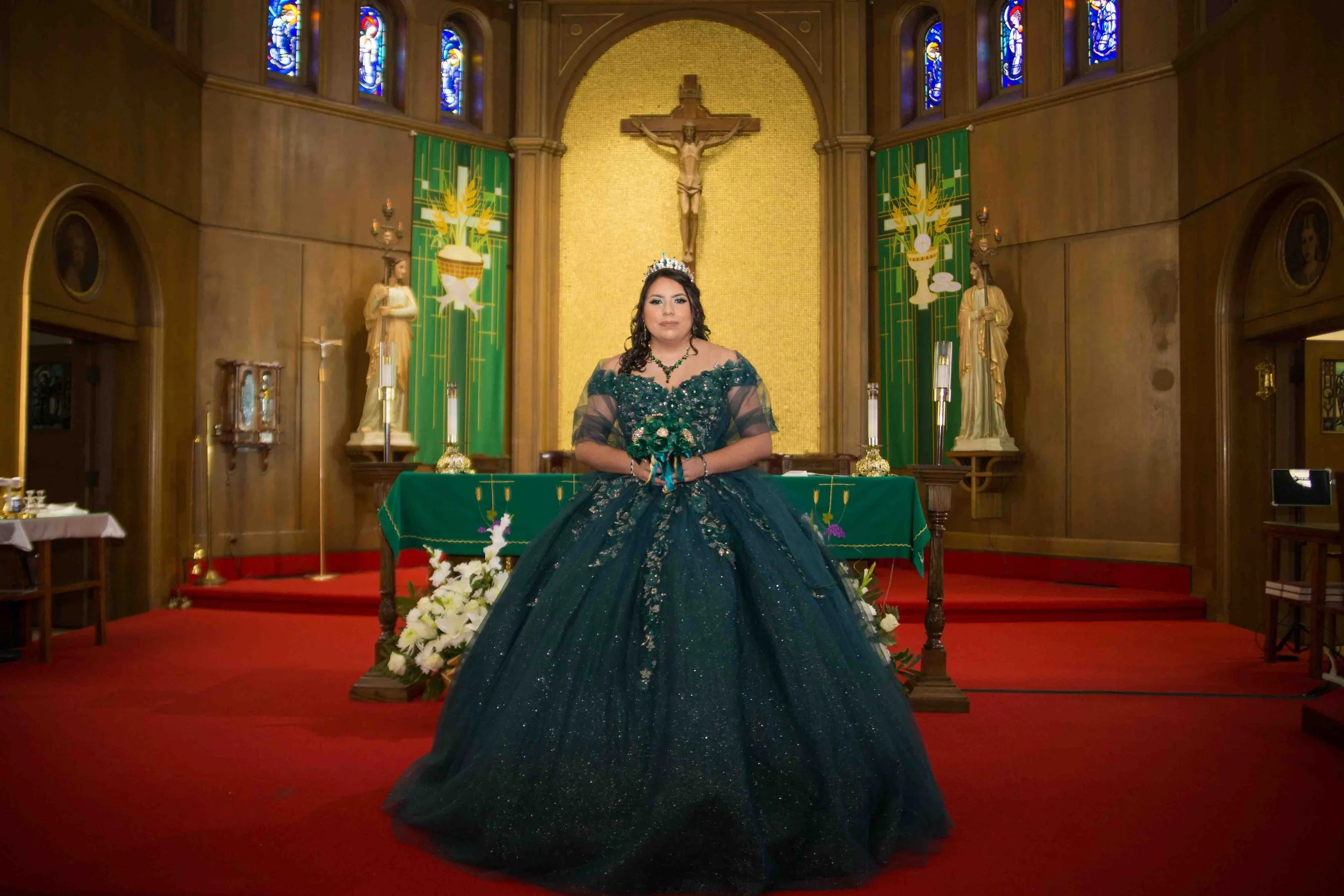 Quinceañera portrait of a young woman in a dark blue glittering ball gown standing at a church altar with a gold crucifix in the background. She wears a tiara, necklace, and gloves, holding a bouquet of flowers, with candles and floral arrangements d
