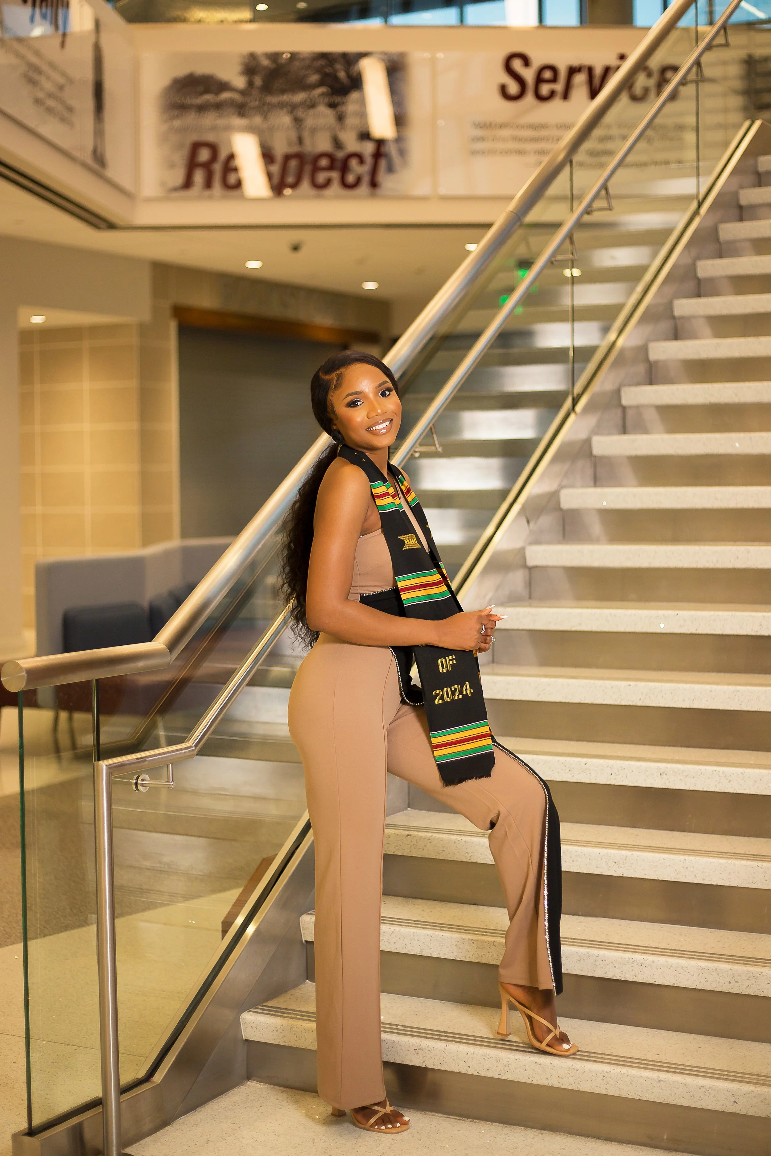 A woman in a tan dress and high heels posing on a staircase, wearing a black stole with colorful stripes that reads 'Class of 2024' at a graduation event.