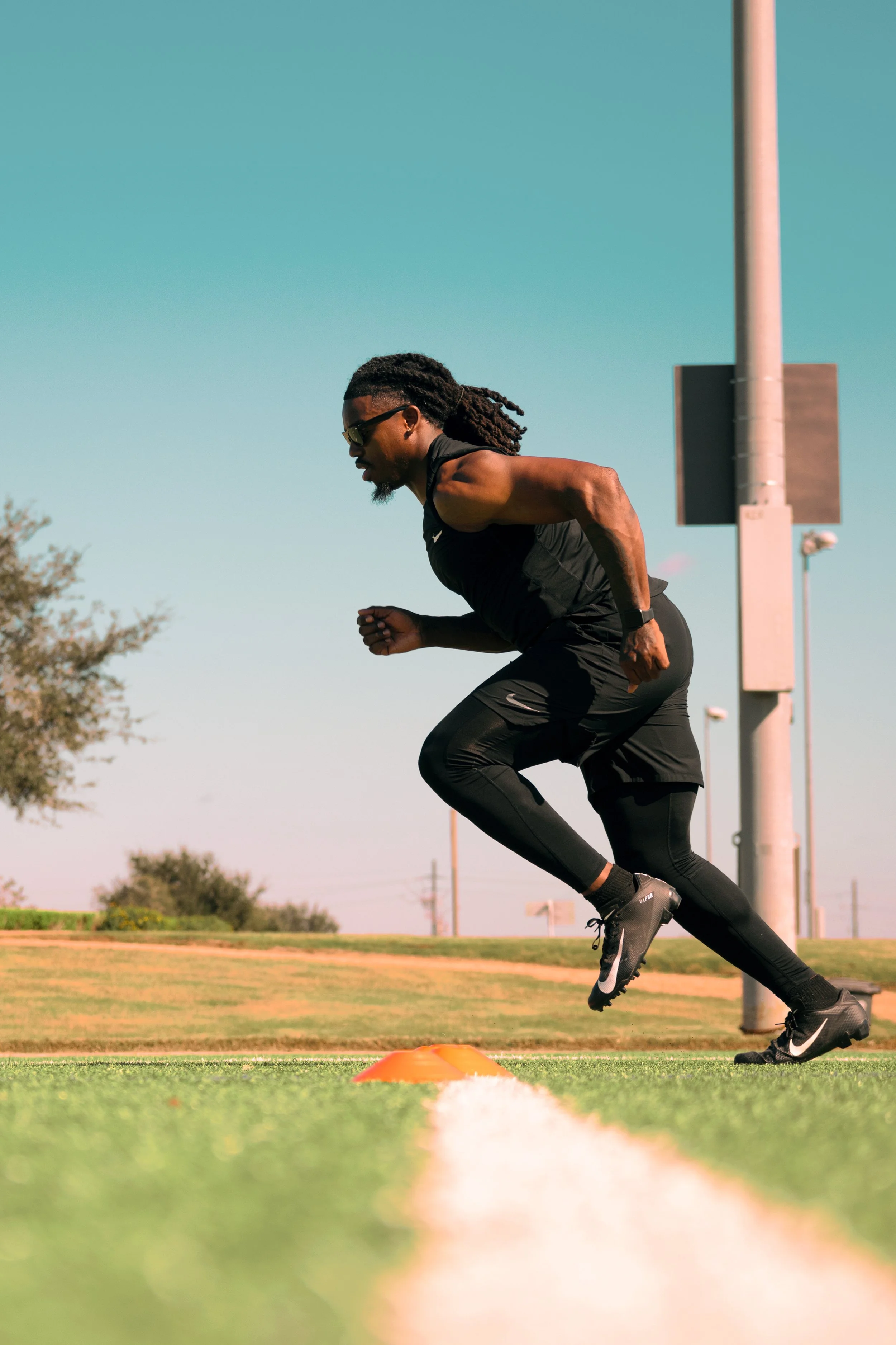 A male athlete in black running gear sprinting on a track with an orange cone nearby, outdoors on a sunny day.