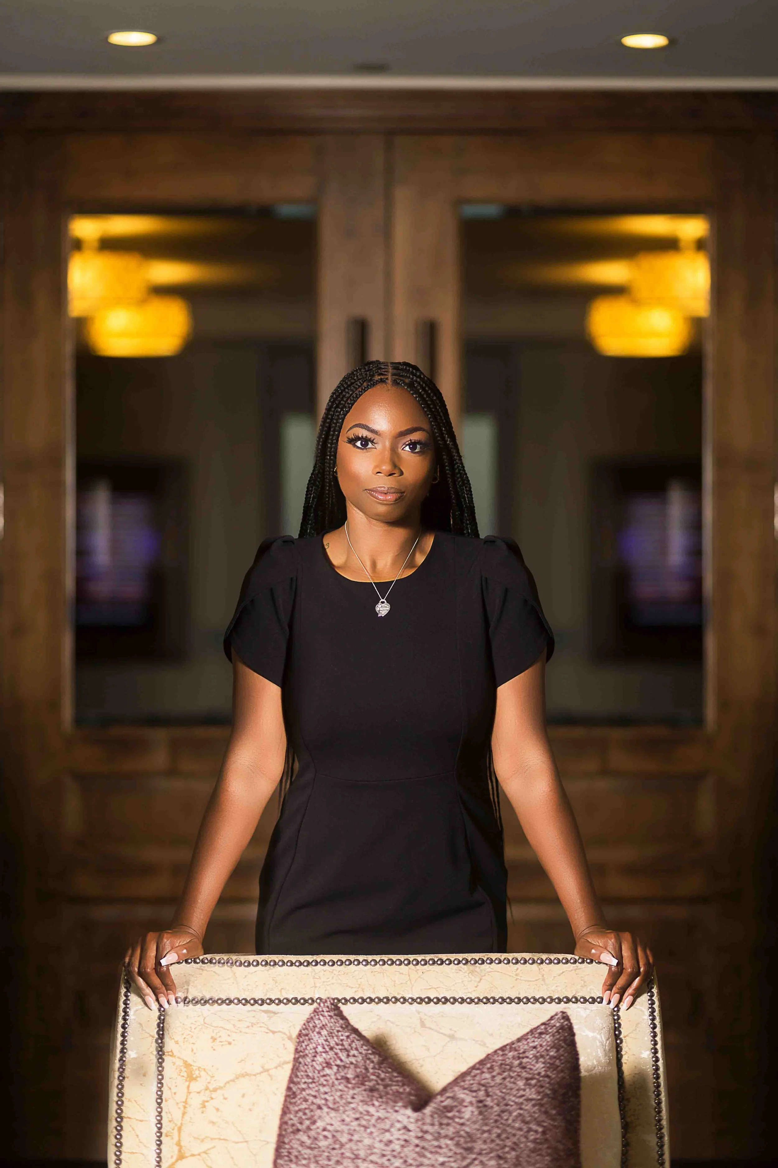 Houston lawyer branding portrait by Lev’s Photography featuring a woman in a black dress standing indoors holding a beige chair in a modern office setting with wooden cabinets and pendant lights