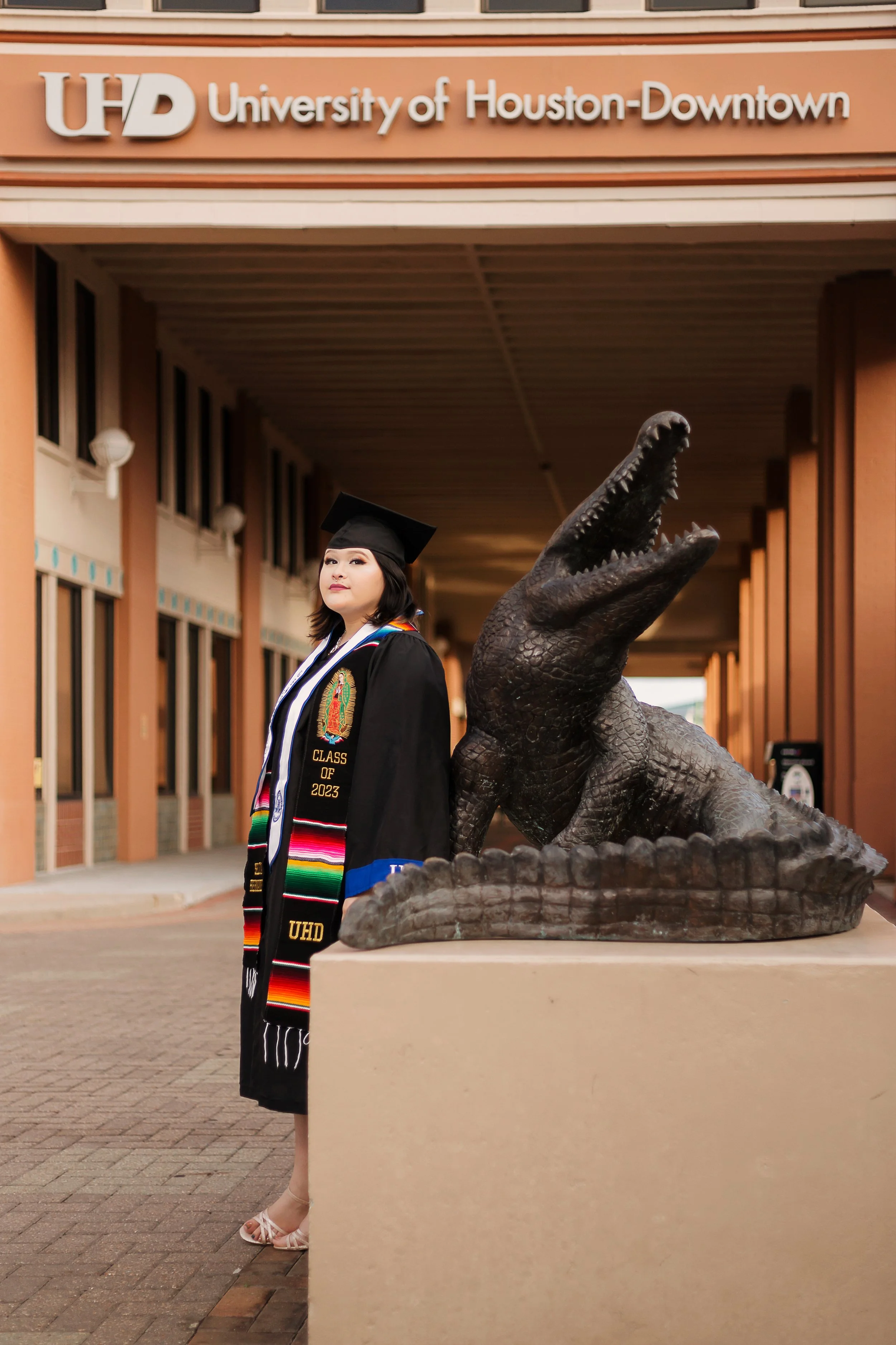 Graduation student in black gown and cap standing next to a crocodile statue in front of the University of Houston-Downtown building.