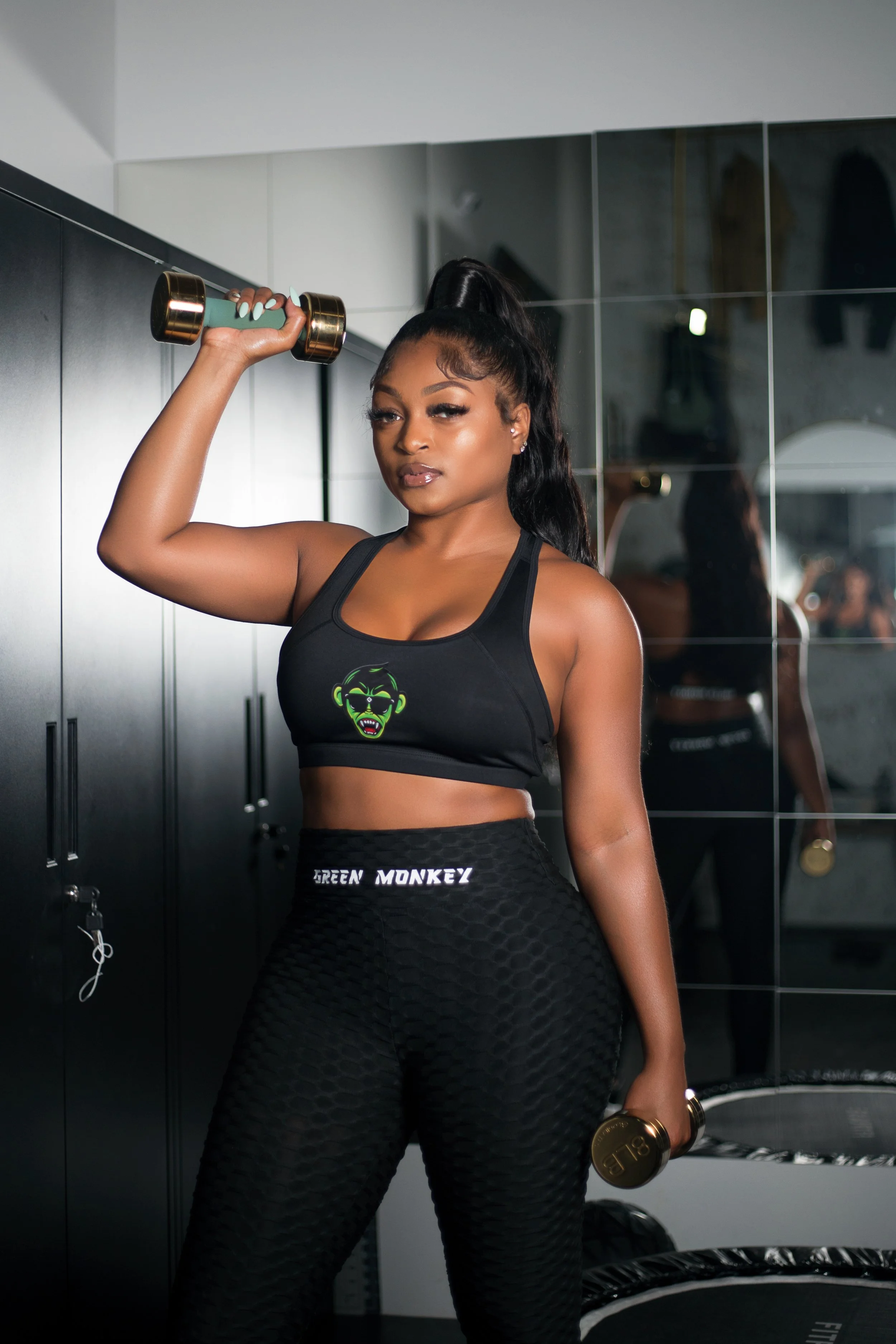 A woman in workout clothes lifting dumbbells in a gym, standing next to black lockers and a mirror.
