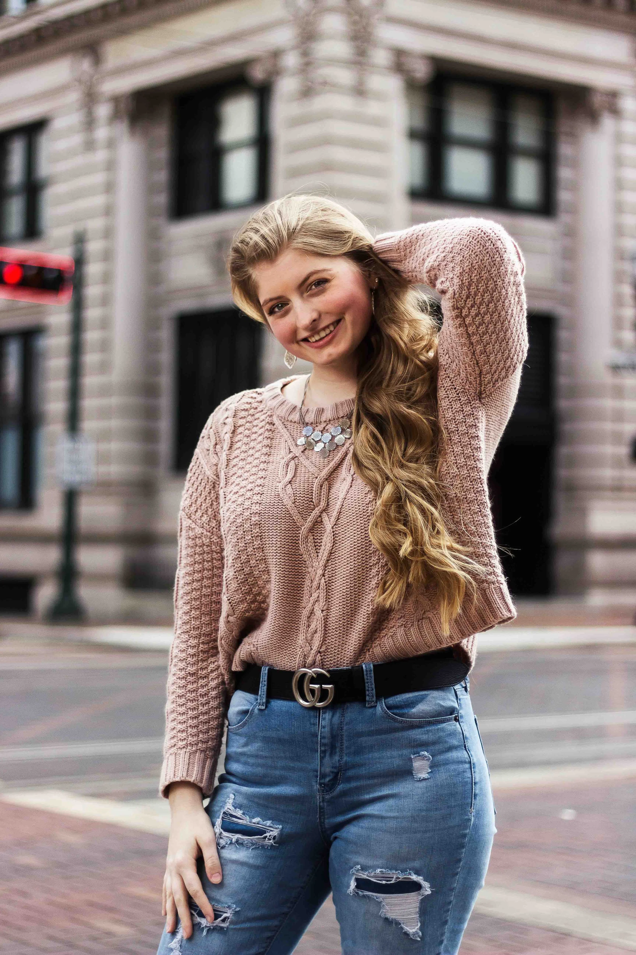 Downtown Houston lifestyle portrait by Lev’s Photography featuring a young woman with long wavy blonde hair wearing a pink knitted sweater, ripped jeans, and statement accessories smiling on a city street