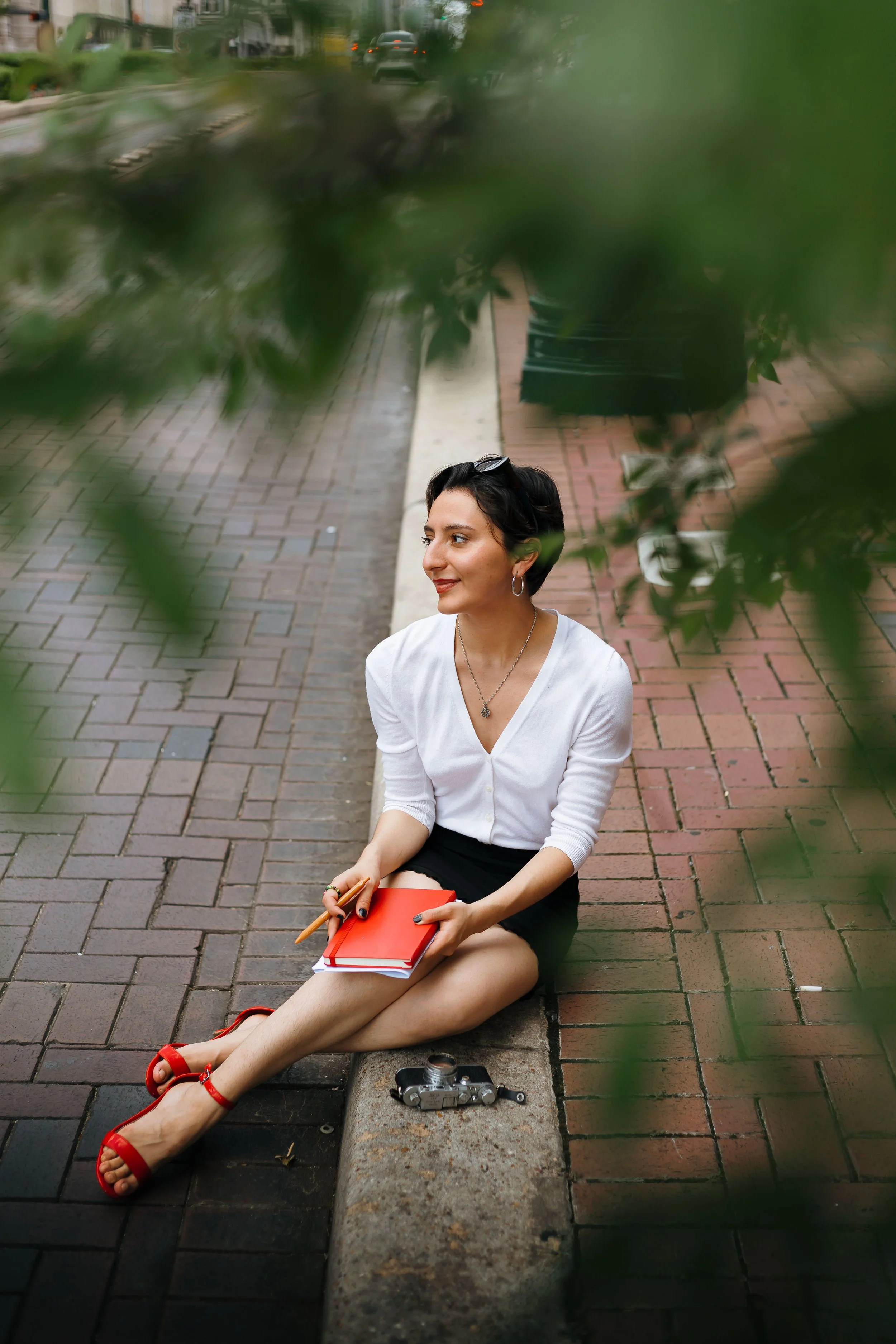Young woman with short dark hair, sitting on a city sidewalk, holding a red notebook and pen, wearing a white blouse, black skirt, red heels, with sunglasses on her head and a camera on the ground nearby, as seen through green leaves.