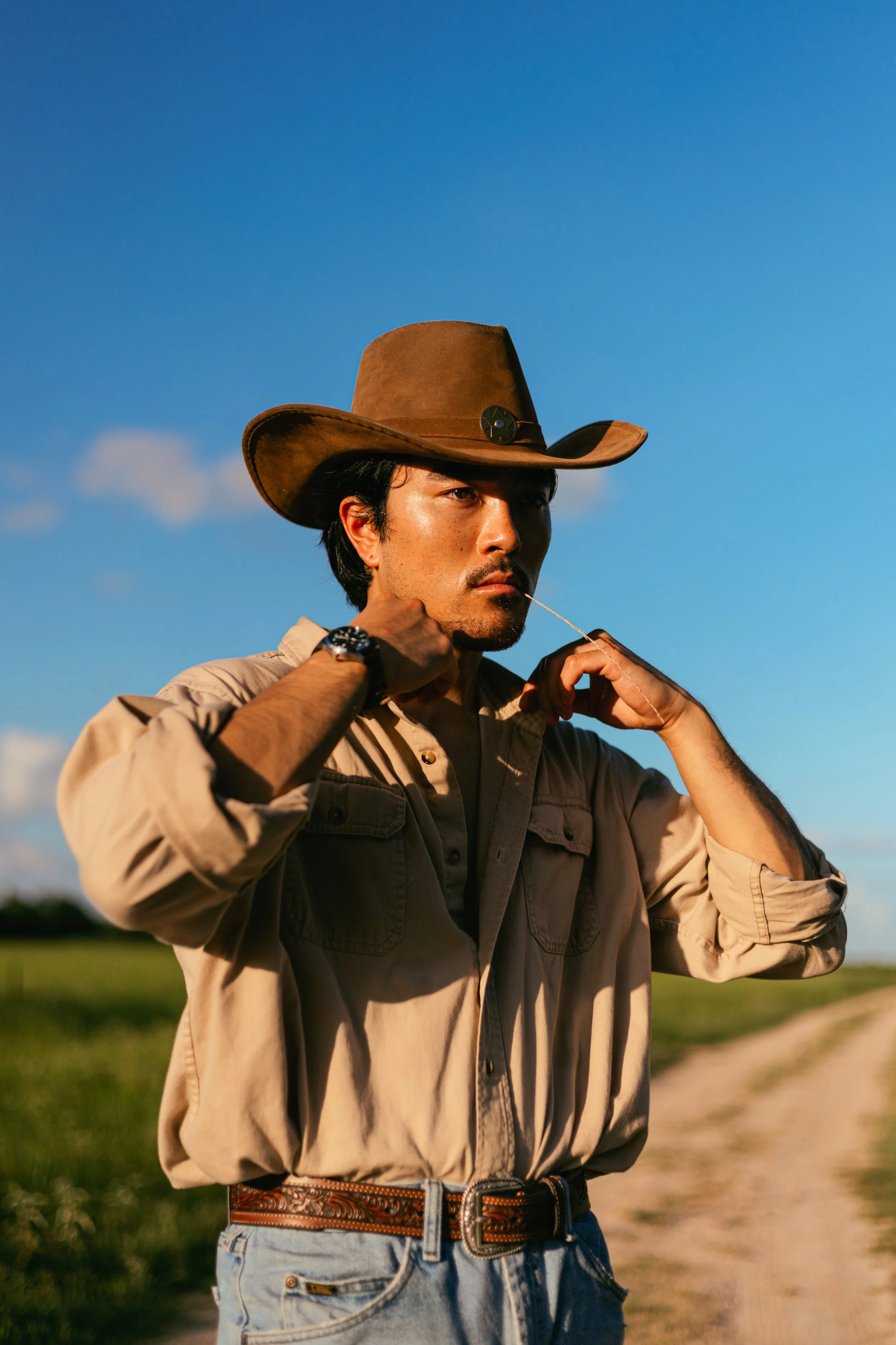 Man in cowboy hat and tan shirt standing on dirt road in countryside with blue sky.