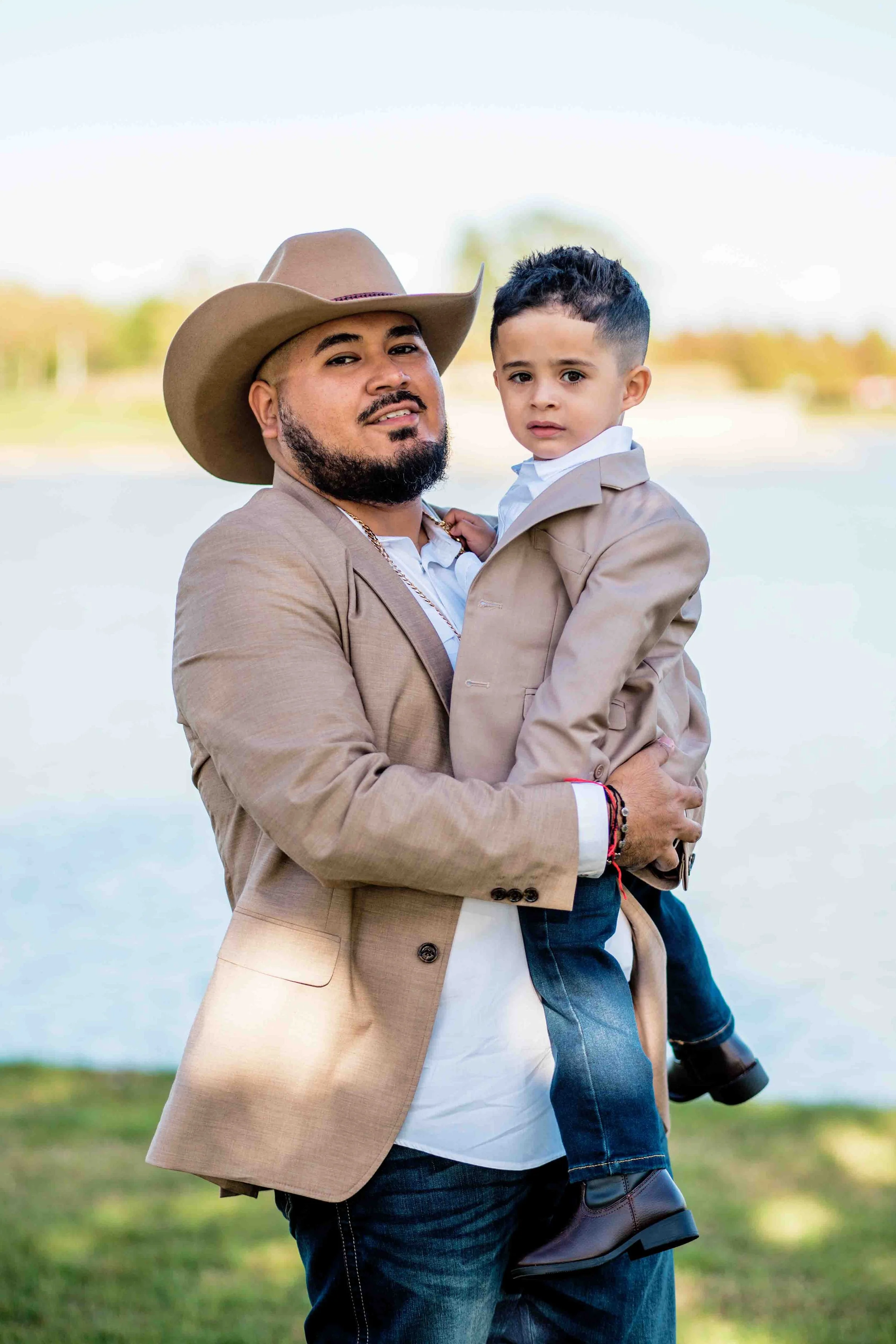 Outdoor family portrait in Houston, Texas by Lev’s Photography featuring a man in a beige suit and cowboy hat holding a young boy in a matching beige suit by a lake with trees and sky in the background
