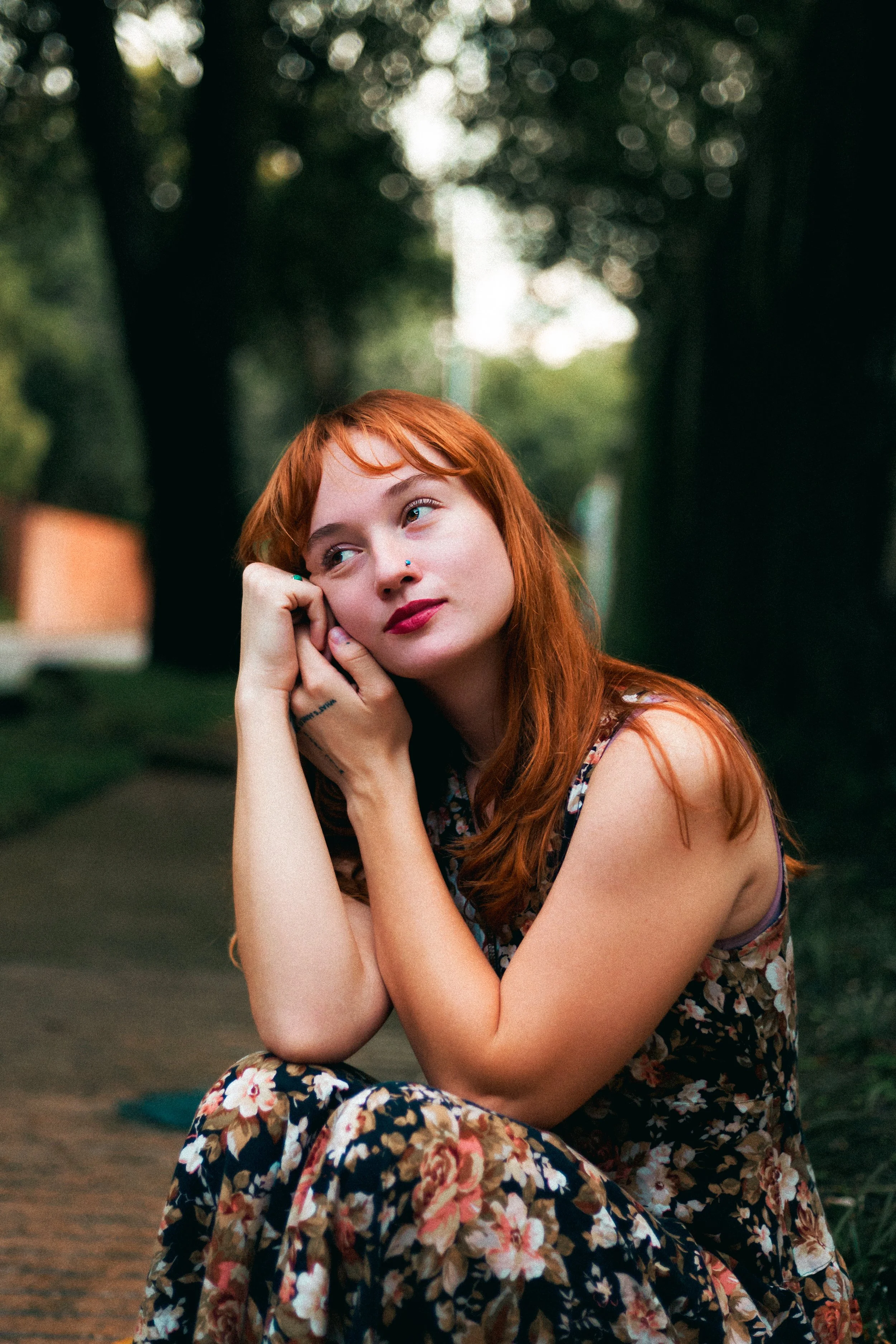 A woman with red hair and a floral dress sitting on a park path, resting her head on her hand and looking thoughtful in a shaded outdoor setting.