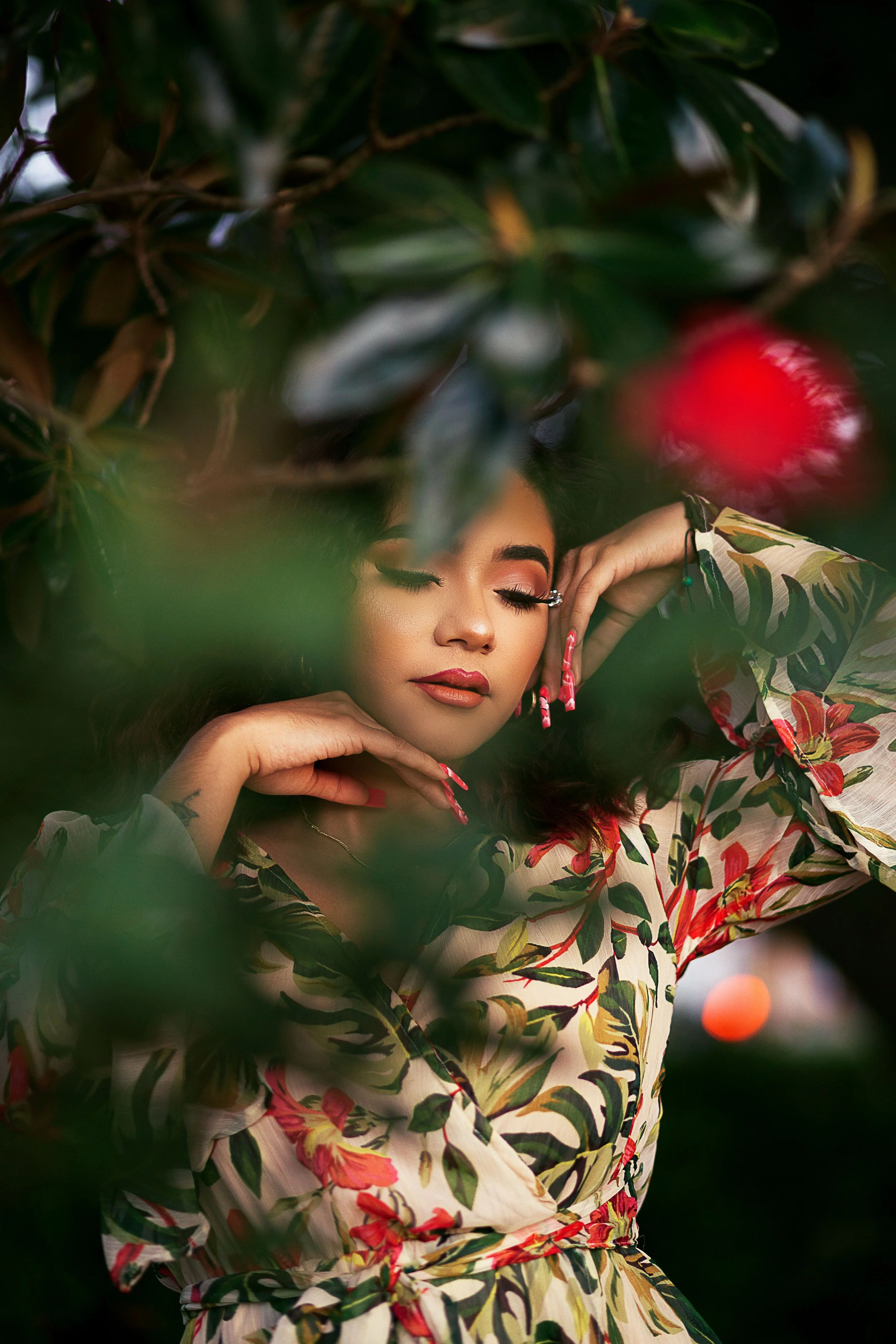 A woman with closed eyes and makeup, wearing a floral-patterned dress, surrounded by green leaves and branches.